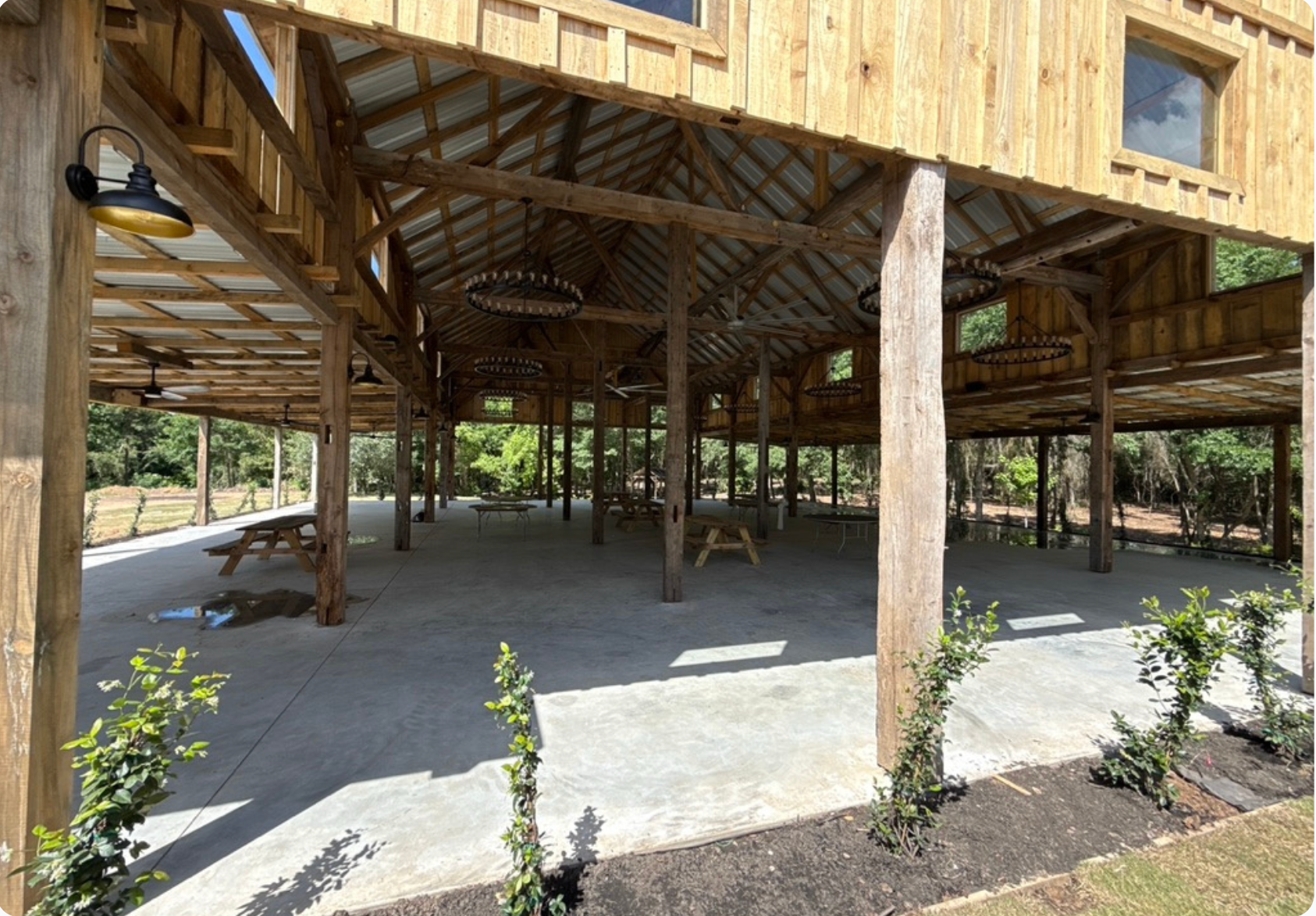 Open-air wooden barn with concrete floor, picnic tables, and landscaping under a metal roof.