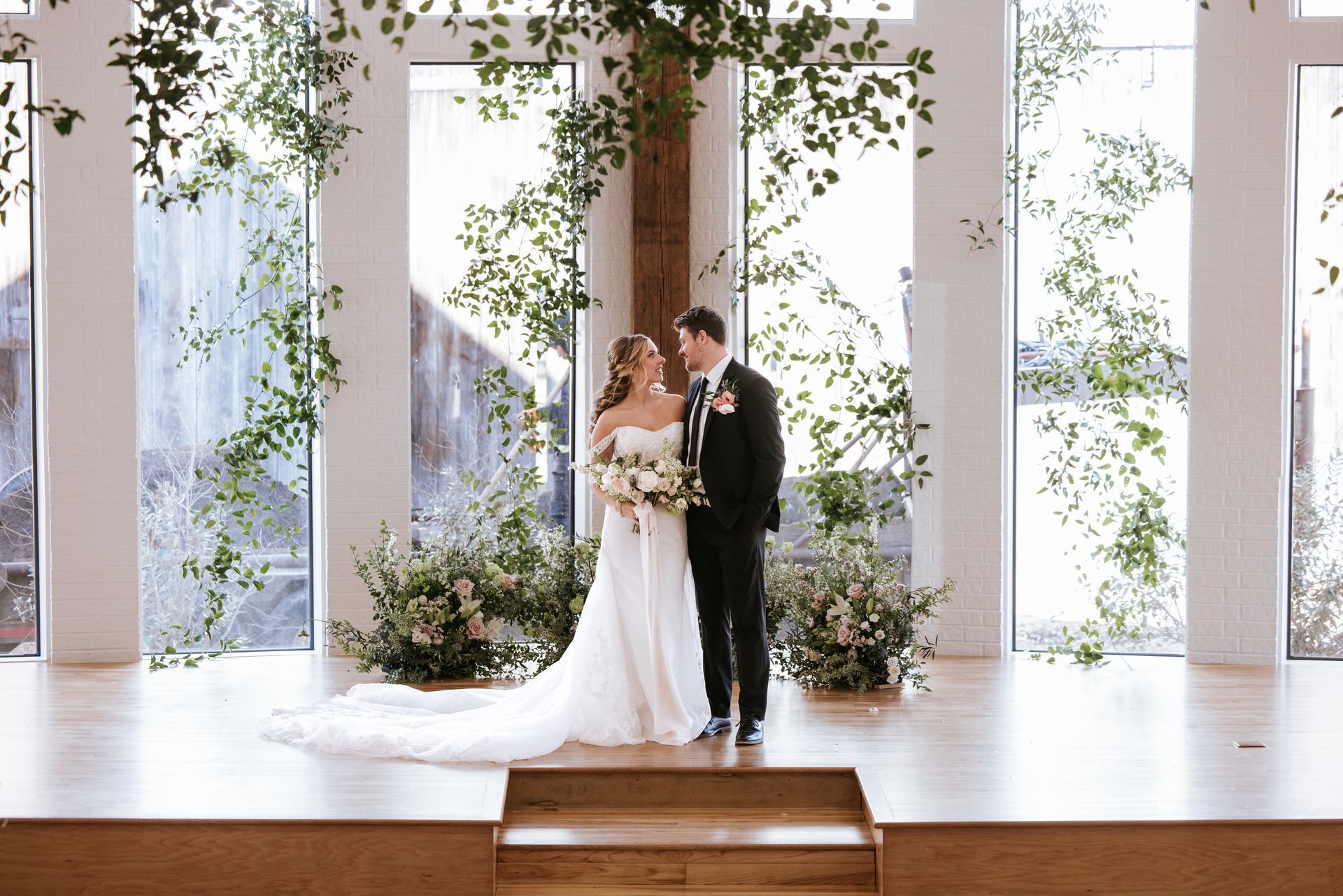 Bride and groom pose on a stage, tall windows behind them, decorated with greenery.