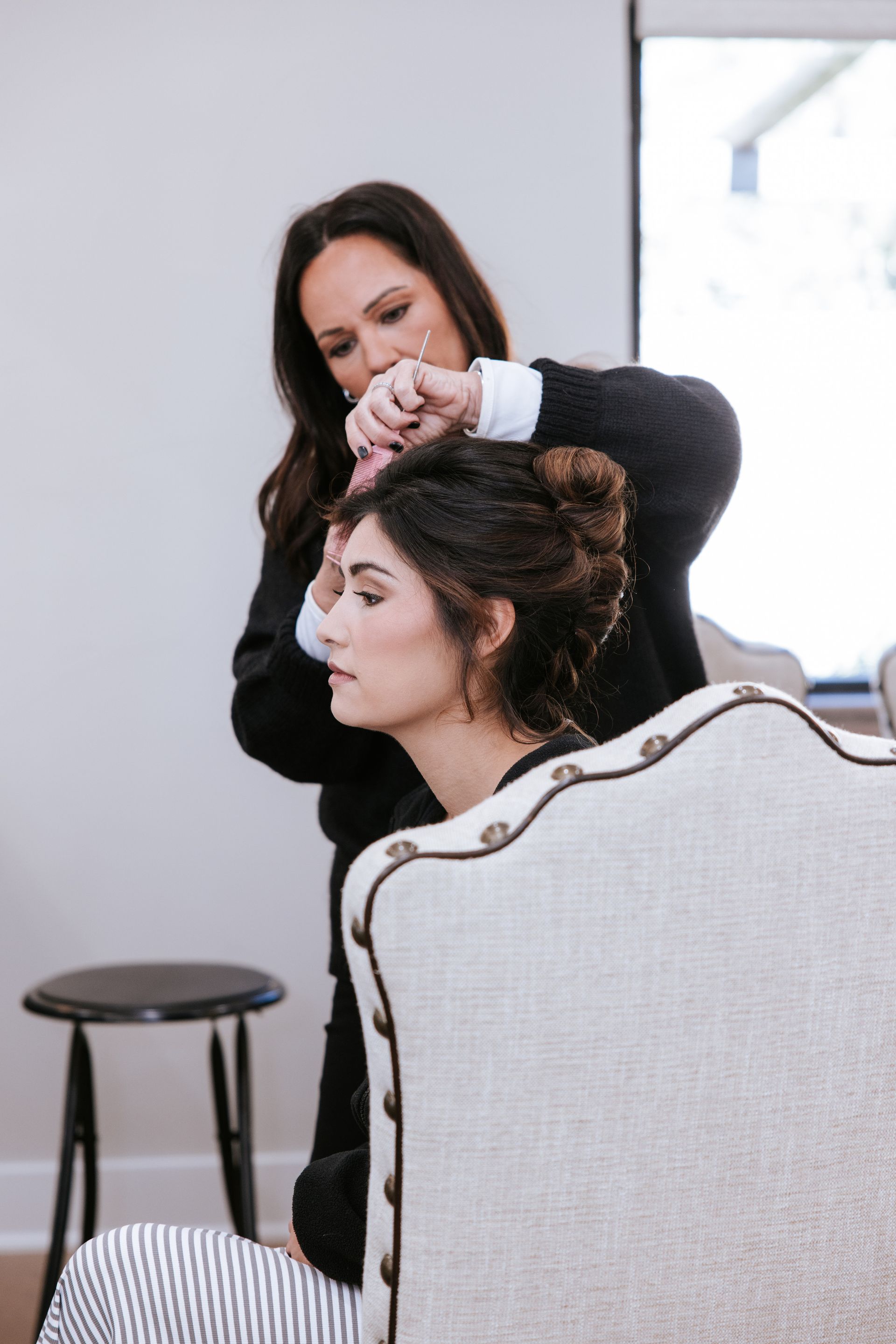 Stylist pinning up a seated client’s hair in a bright salon, with a stool and upholstered chair nearby