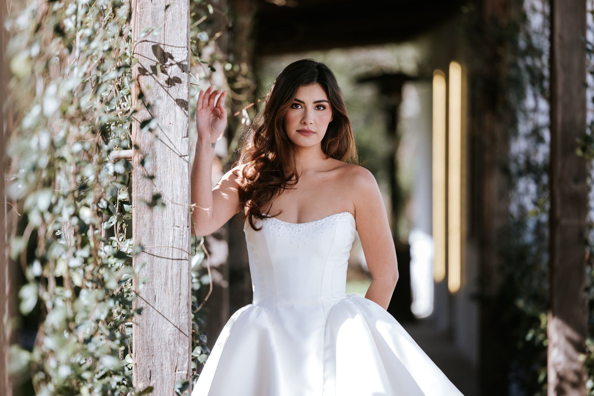 Bride in a white strapless wedding gown standing by ivy-covered columns in a covered walkway