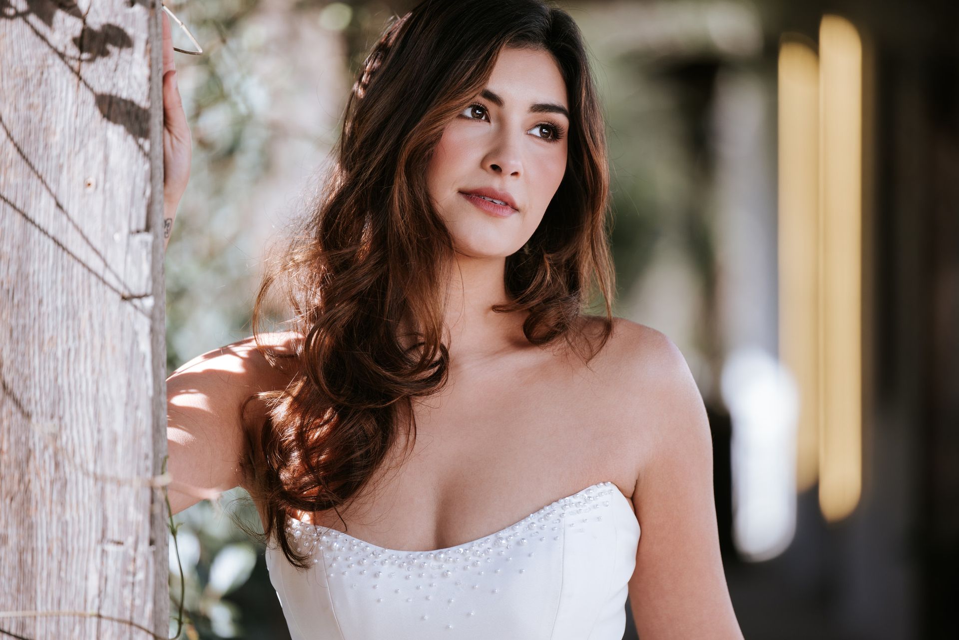 Woman in a white strapless dress standing by a brick wall outdoors, looking to the side.