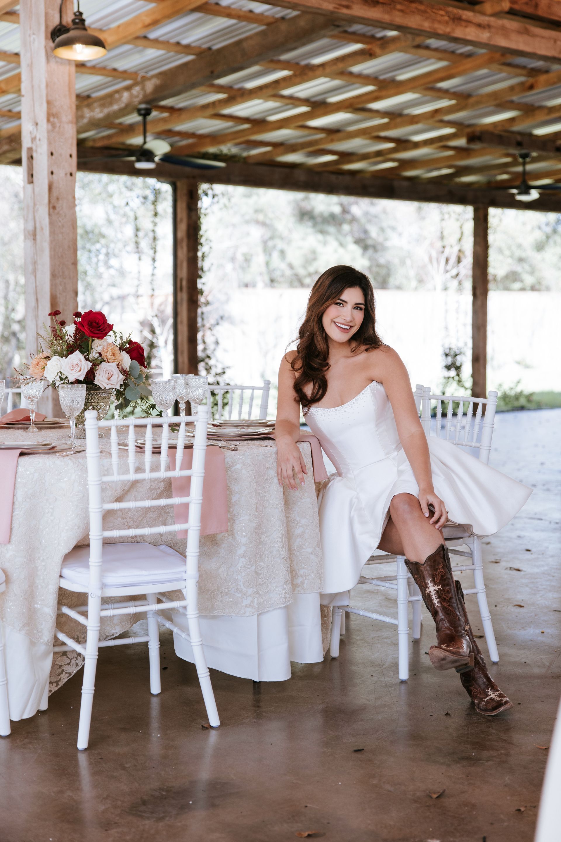 Woman in white dress and brown boots posing at a decorated outdoor reception table under a wooden canopy