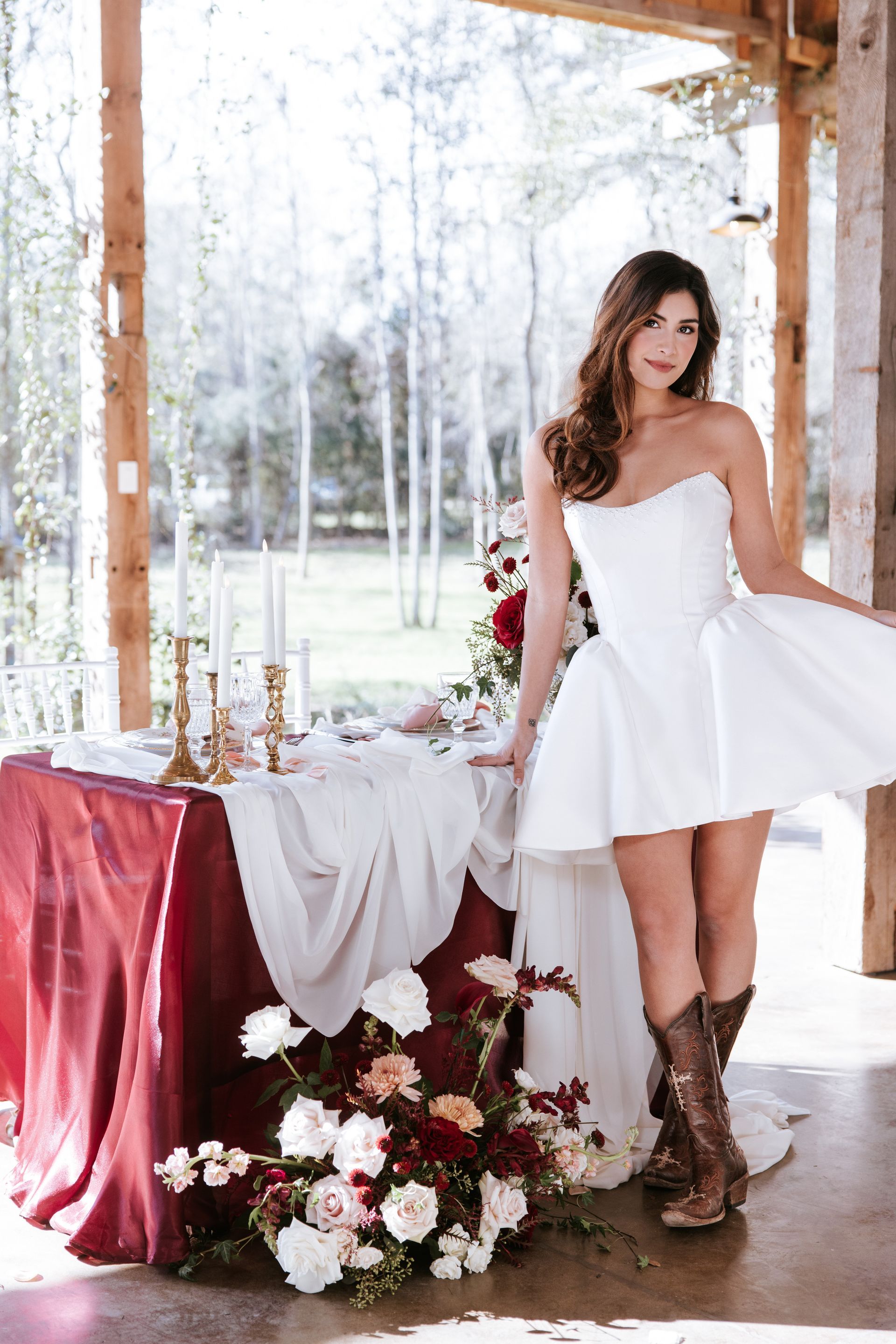 Woman in a white dress and brown boots posing beside a floral table in a rustic, windowed pavilion.