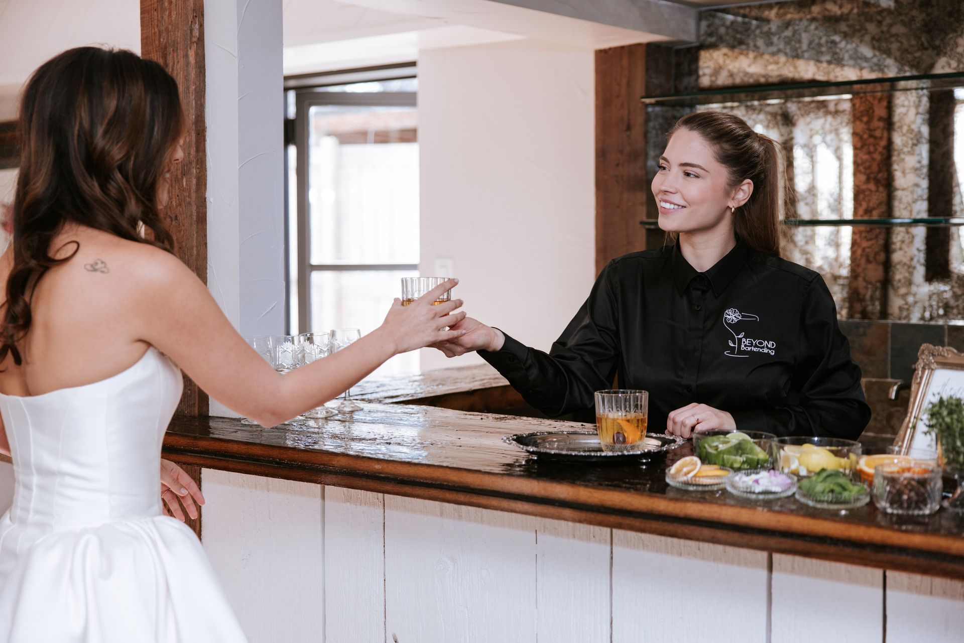 Woman in a white dress hands a glass to a smiling bartender behind a marble bar.