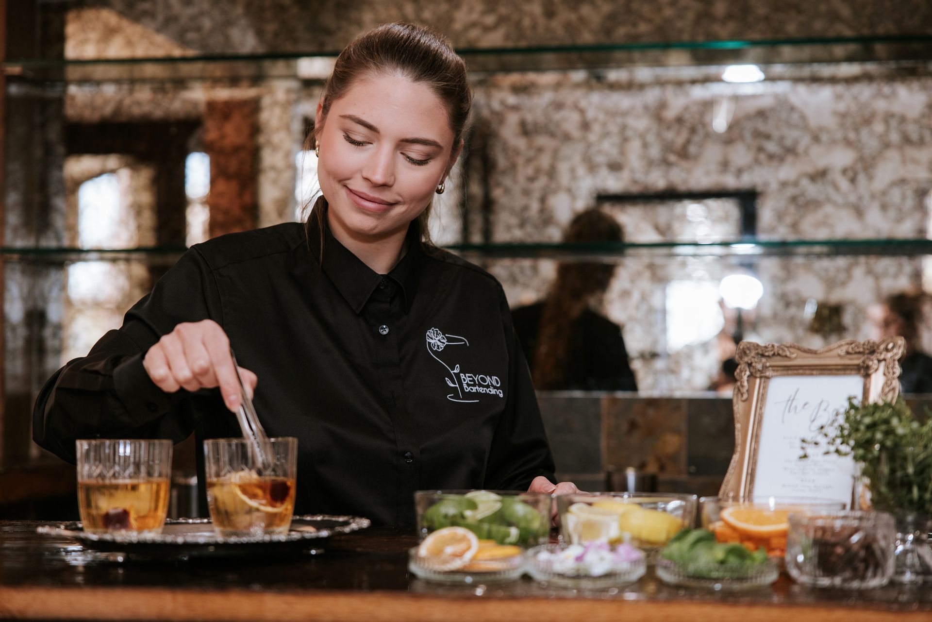 Bartender mixing drinks behind a bar with citrus, herbs, and glassware displayed.