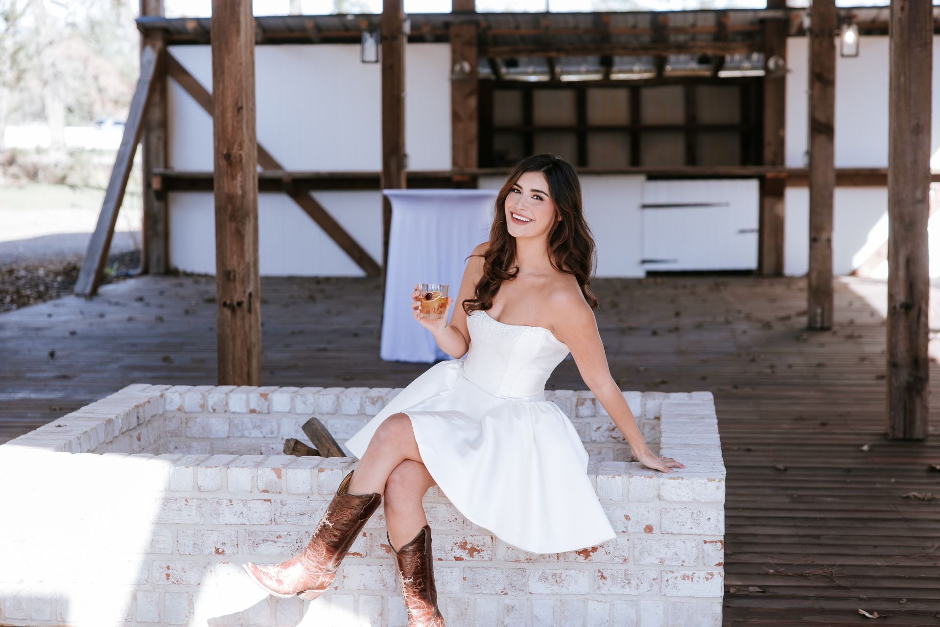 Woman in a white dress sitting on a white brick bench, smiling and holding a drink on a wooden deck.