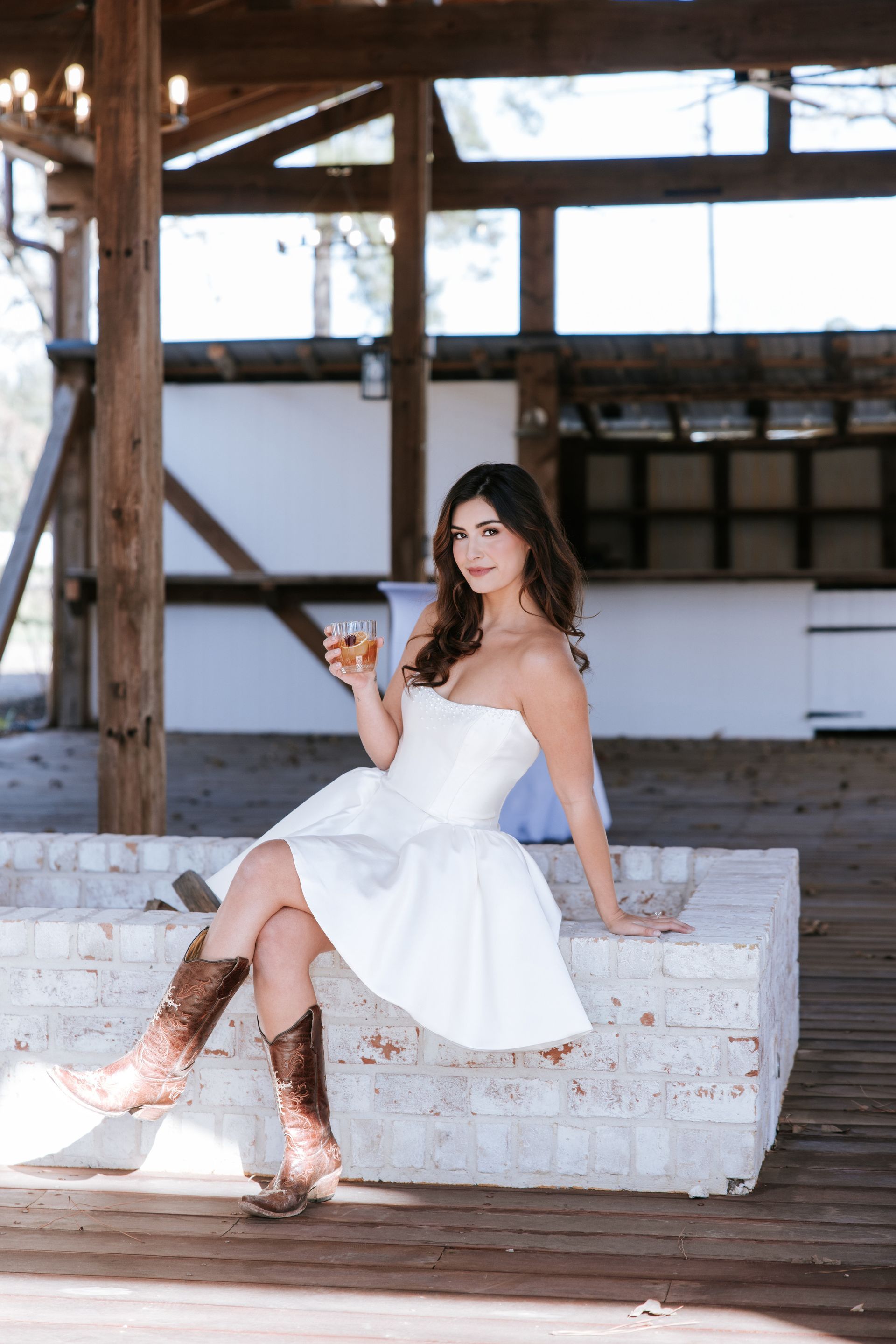 Woman in a white dress sitting on a concrete block in an open rustic warehouse, holding a drink.