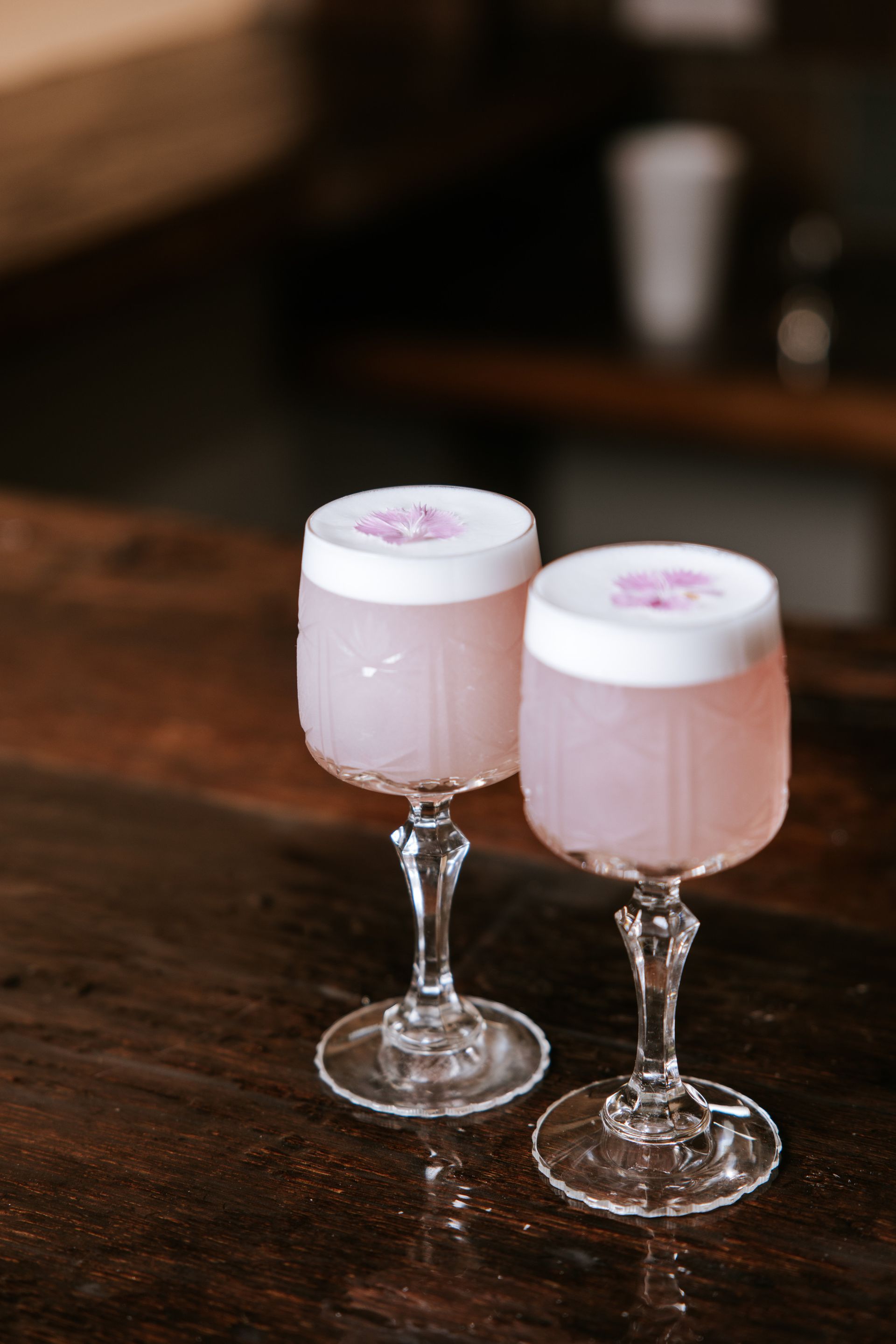 Two pale pink cocktails in stemmed glasses on a wooden table, each topped with frothy white foam and a flower garnish