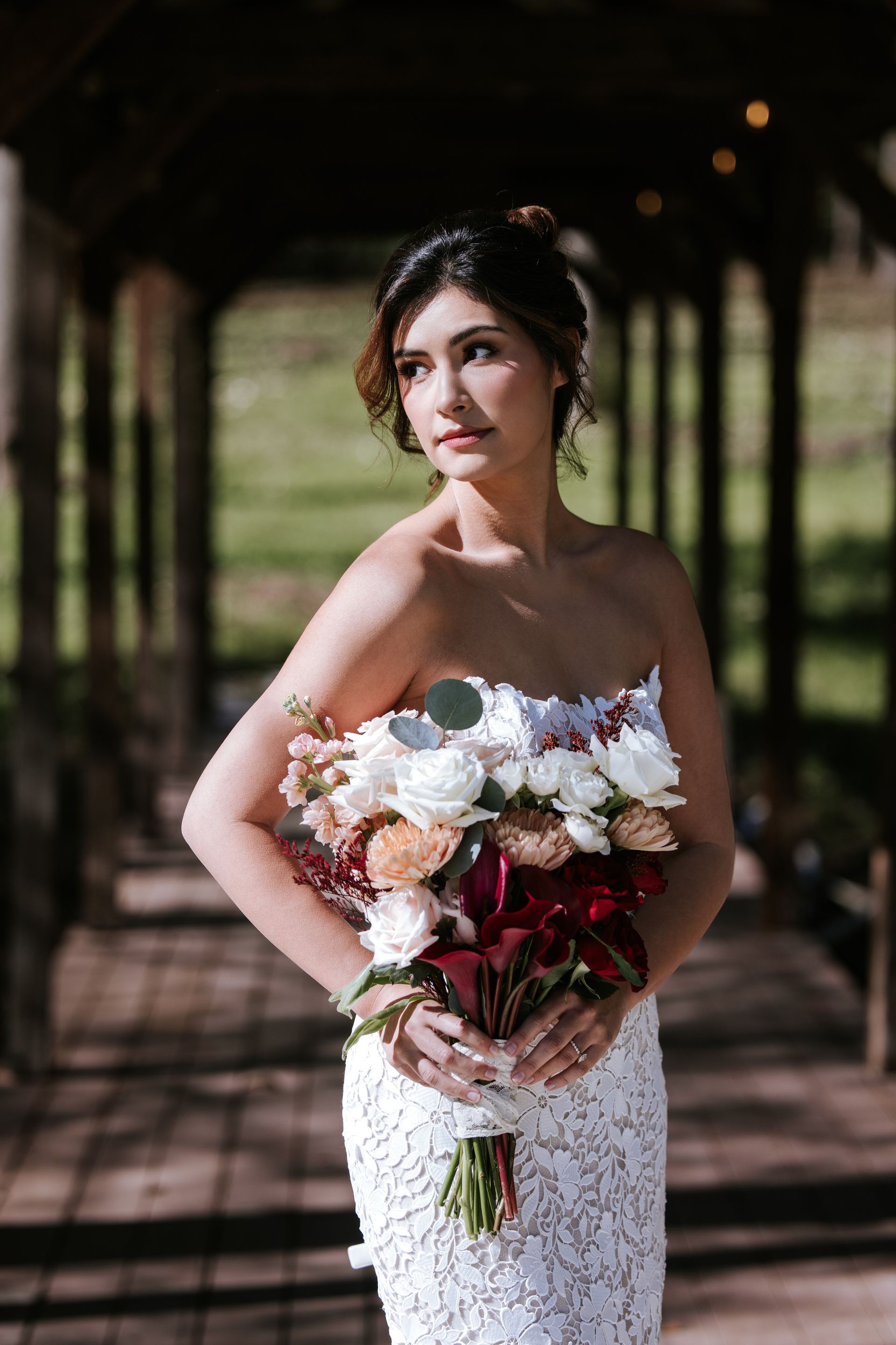 Bride in a white lace gown holding a colorful bouquet in a shaded garden walkway