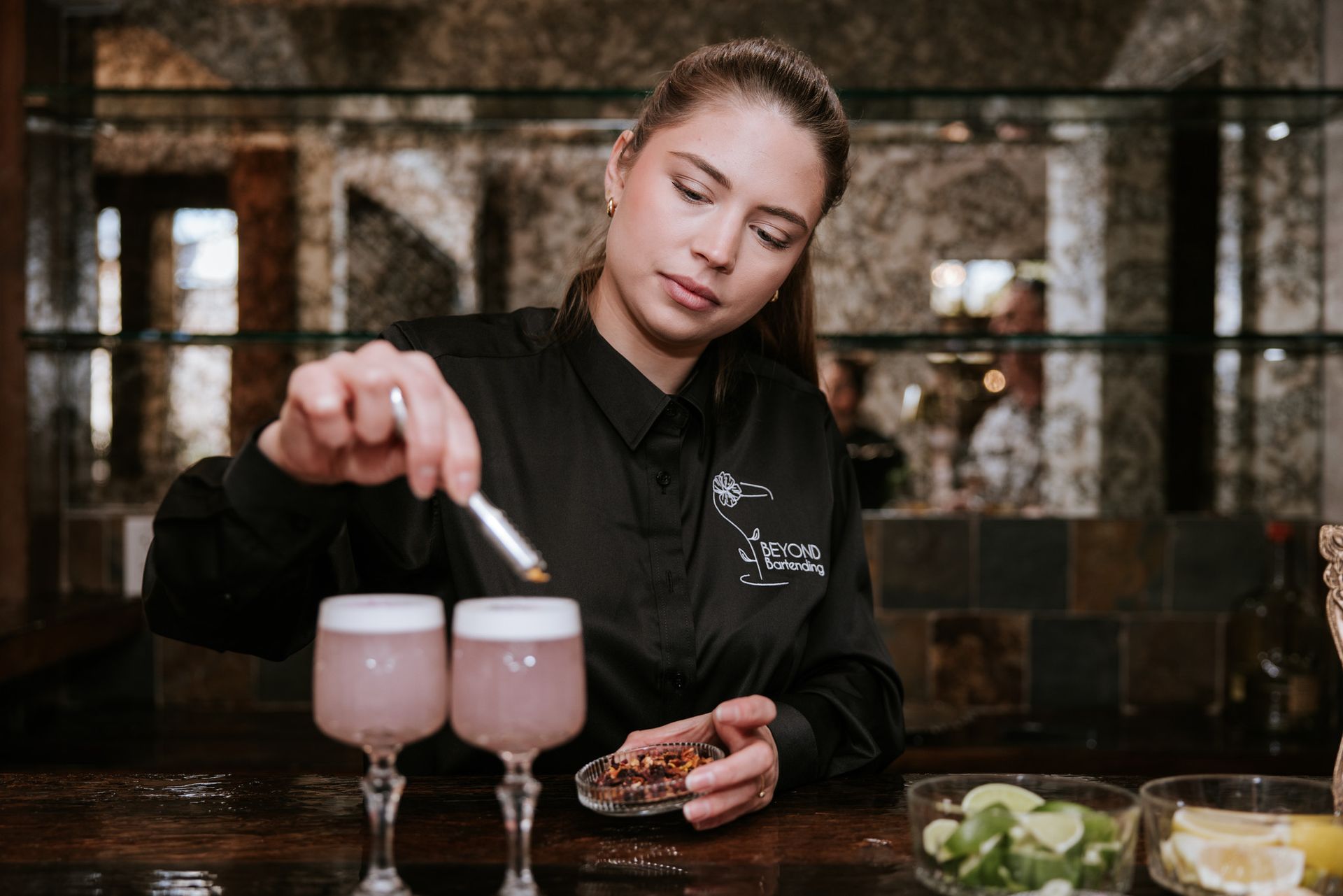 Bartender pouring a drink into a pink cocktail at a bar, with snacks and ingredients nearby