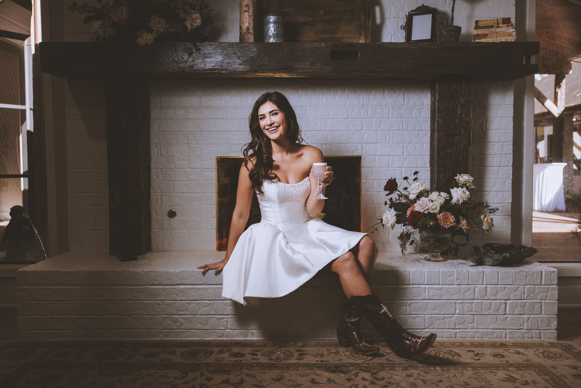 Woman in a white dress sitting by a stone fireplace in a rustic room, smiling with boots on.