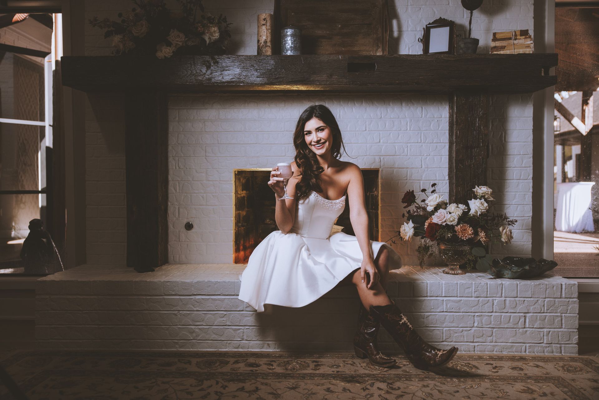Woman in a white dress seated by a fireplace in a cozy, rustic room, holding a small drink and smiling
