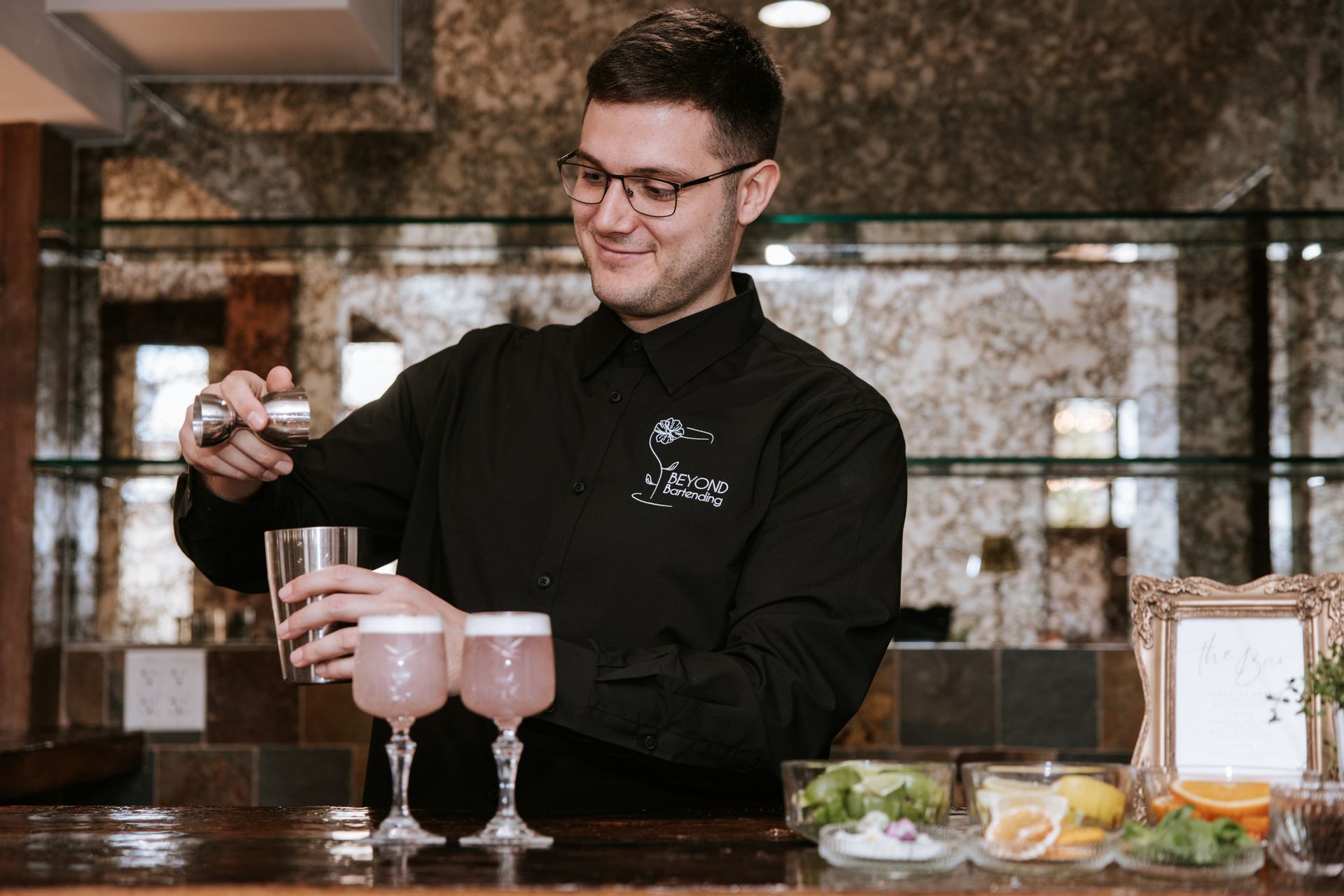 Bartender in black shirt pouring a drink into two pink cocktails at a bar counter