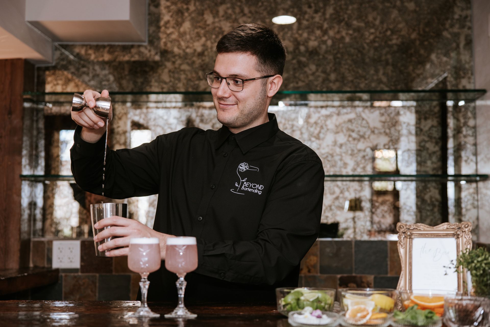 Bartender pouring a cocktail in a stylish bar, with two pink drinks on the counter.