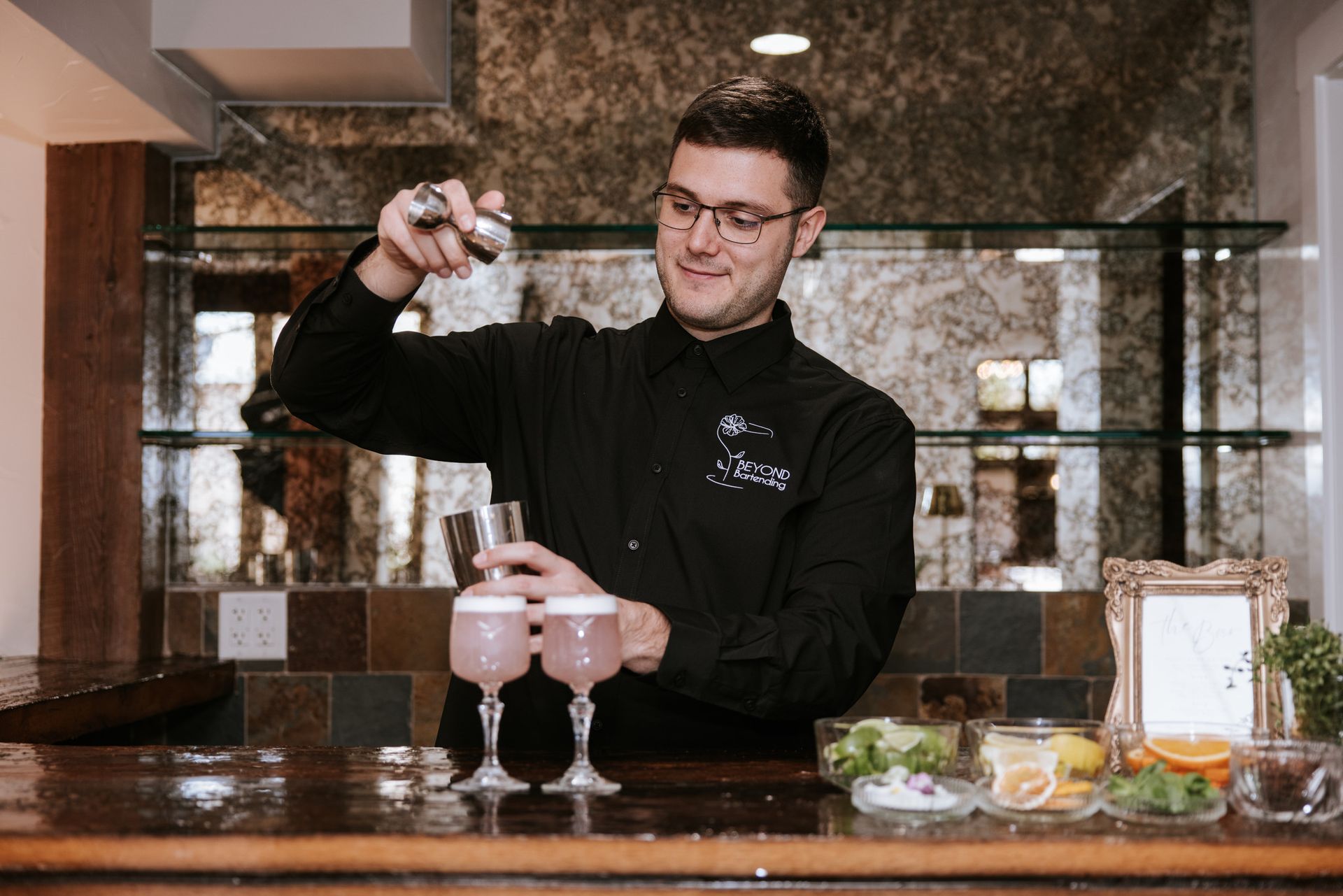 Bartender pouring a drink into a martini glass at a bar, with two cocktails on the counter.