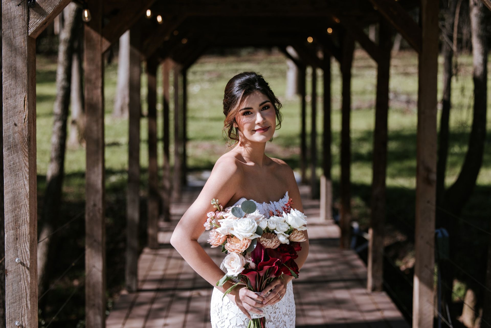 Bride holding a bouquet, standing in a wooden pavilion with a soft smile.