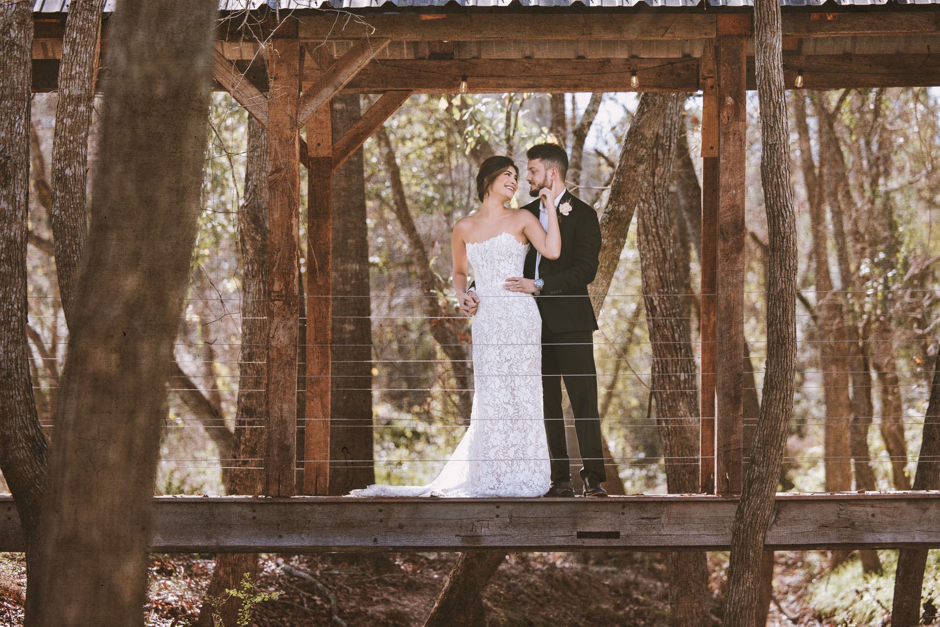 Couple in wedding attire embracing on a rustic wooden bridge in a wooded setting