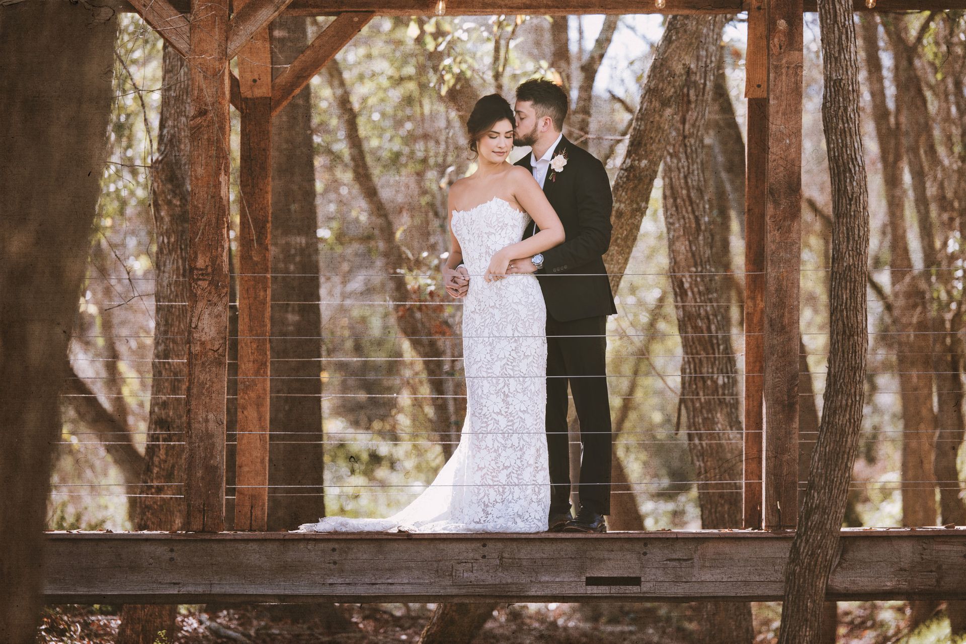 Couple in wedding attire embracing on a wooden bridge in a wooded setting
