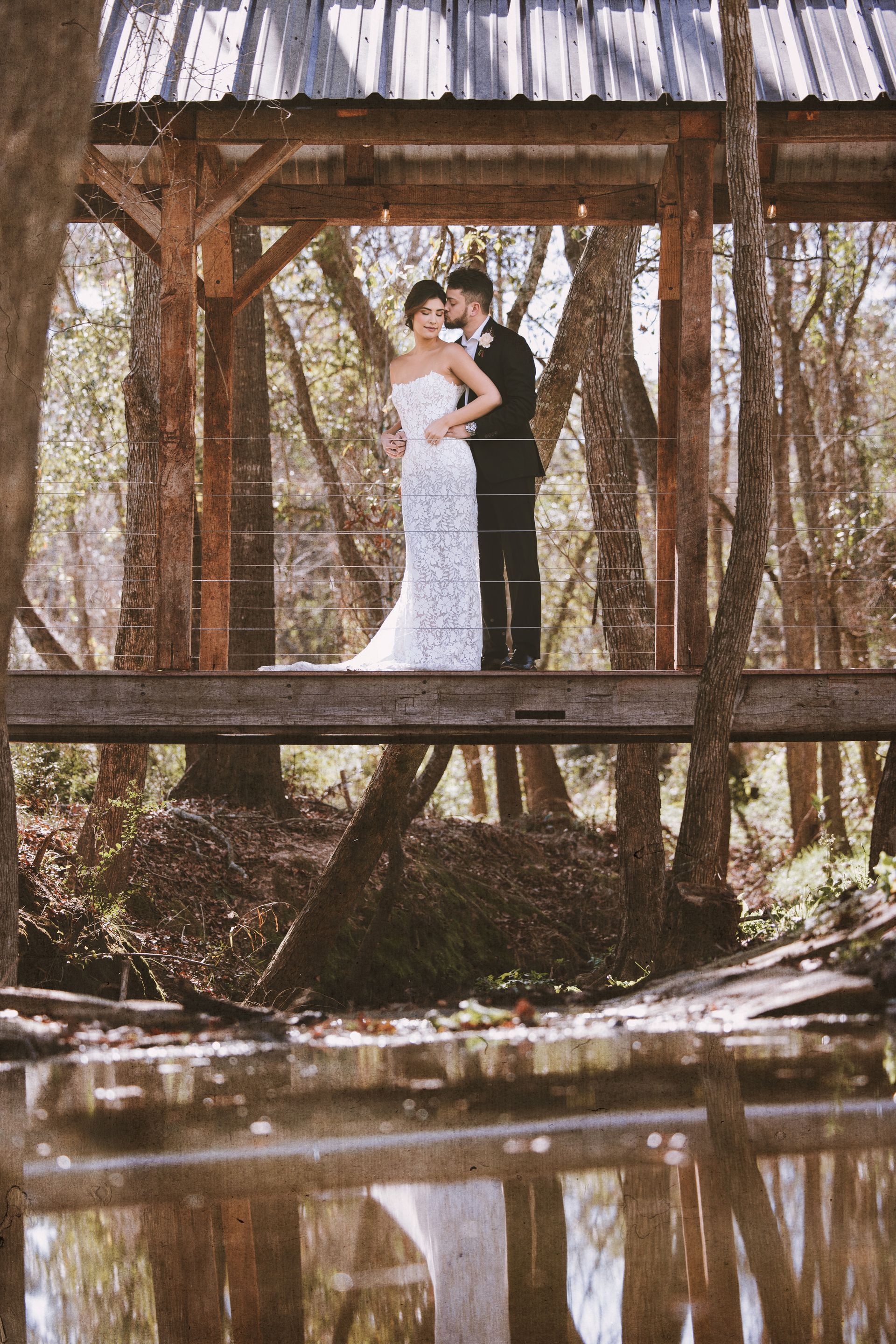 Couple embracing on a rustic wooden bridge over a calm creek, reflected in the water.