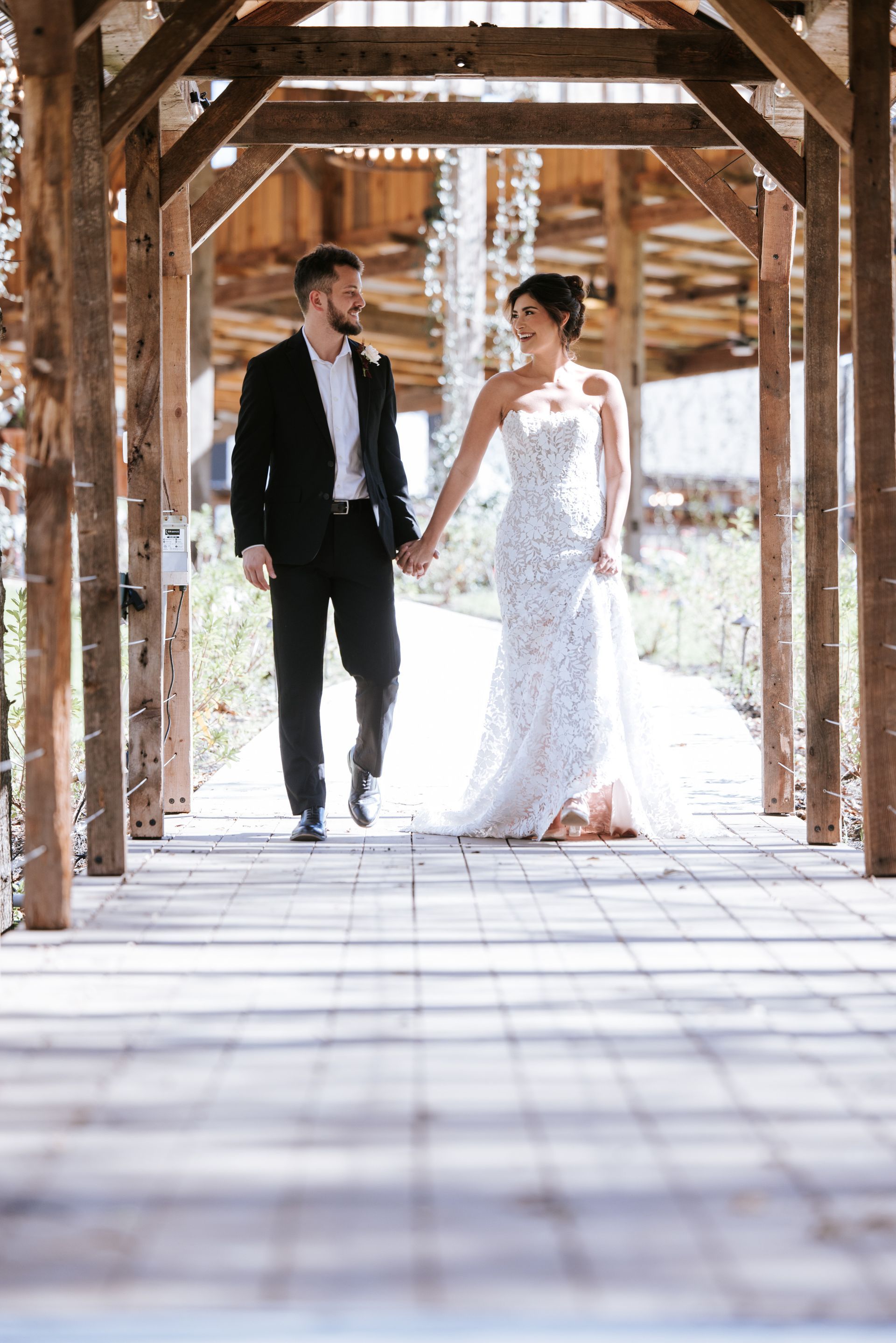Bride and groom holding hands on a wooden bridge in a sunlit rustic pavilion