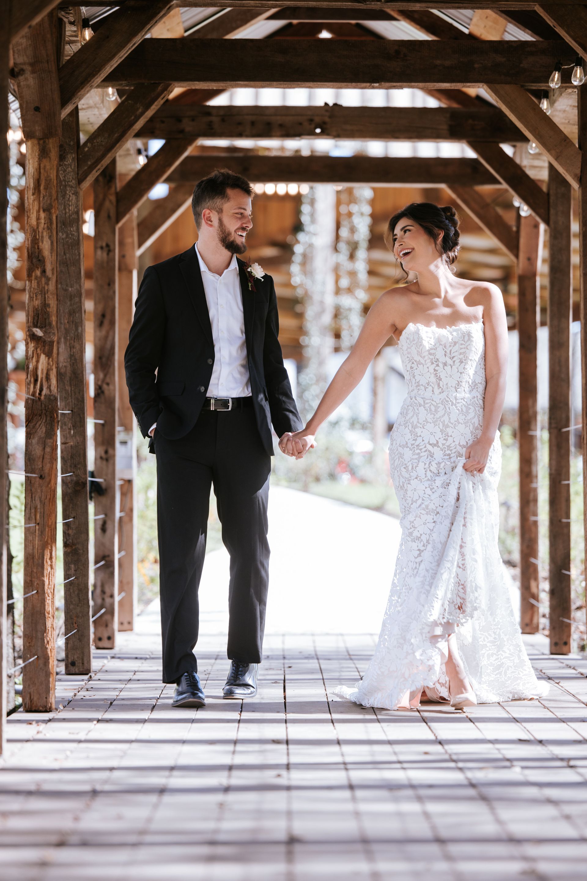 Couple holding hands under a wooden pergola, bride in white lace gown and groom in black suit, outdoors