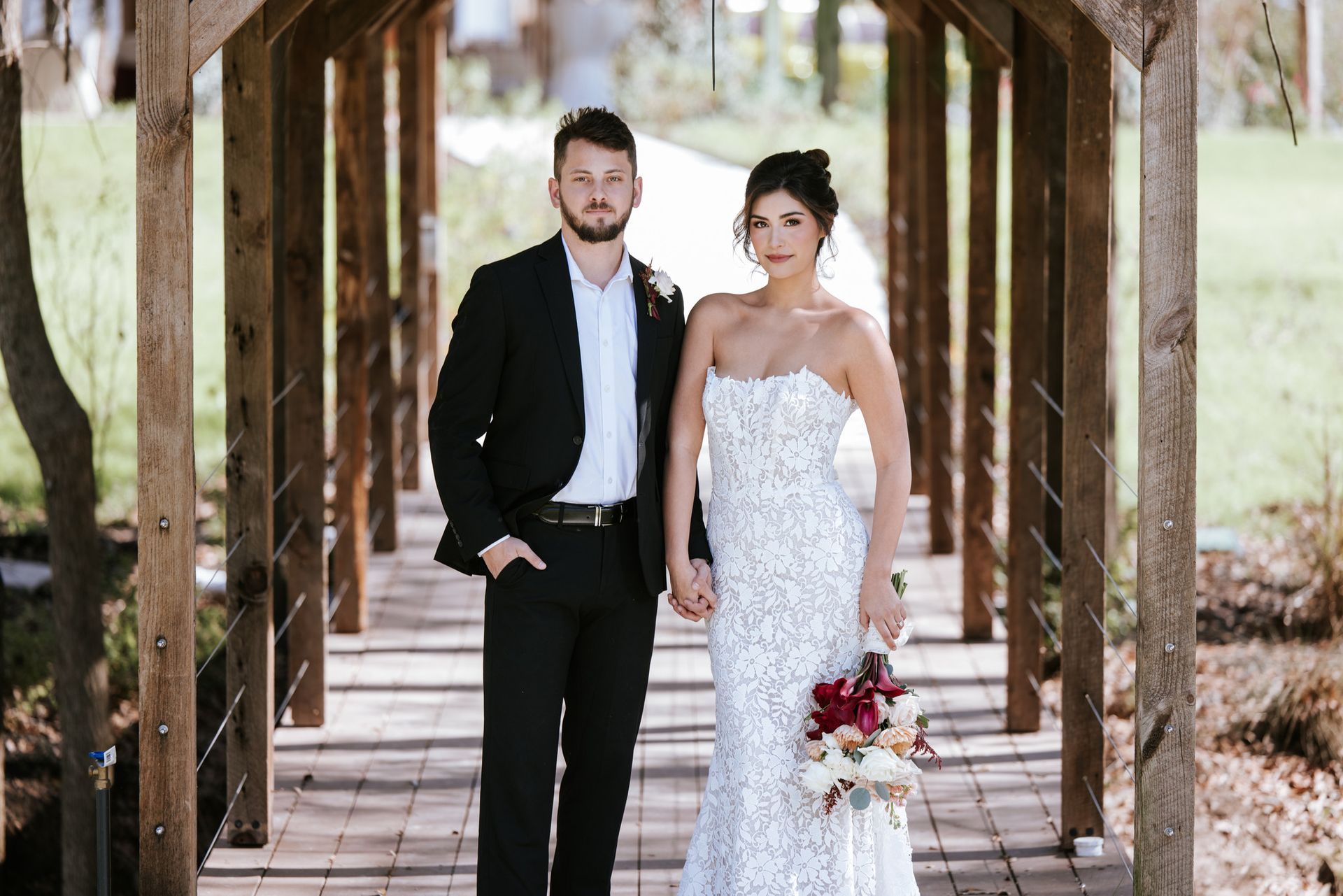 Bride and groom stand on a wooden bridge, the bride holding a red bouquet in a white lace gown.