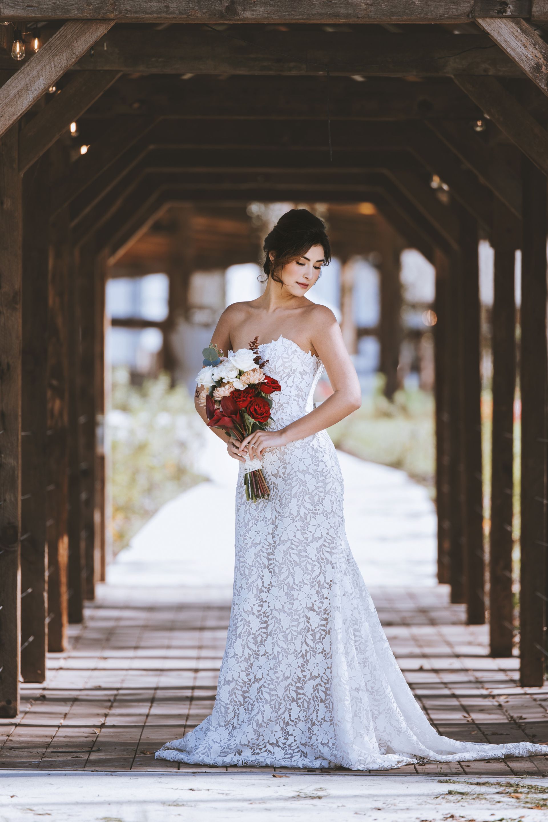 Bride in a white lace gown holding a red-and-white bouquet under a wooden pergola
