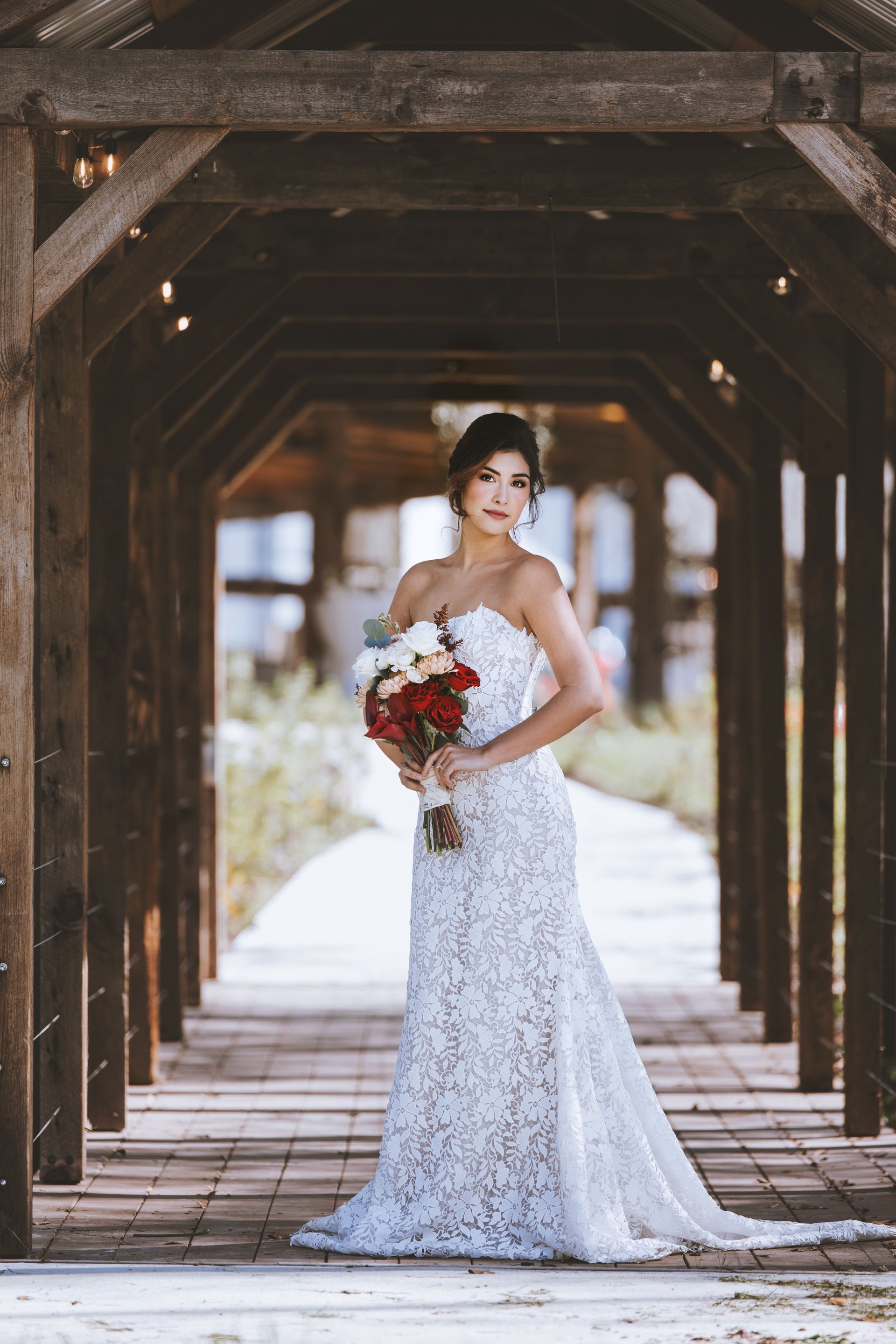 Bride in a white lace gown holding a red bouquet, standing in a wooden covered walkway.