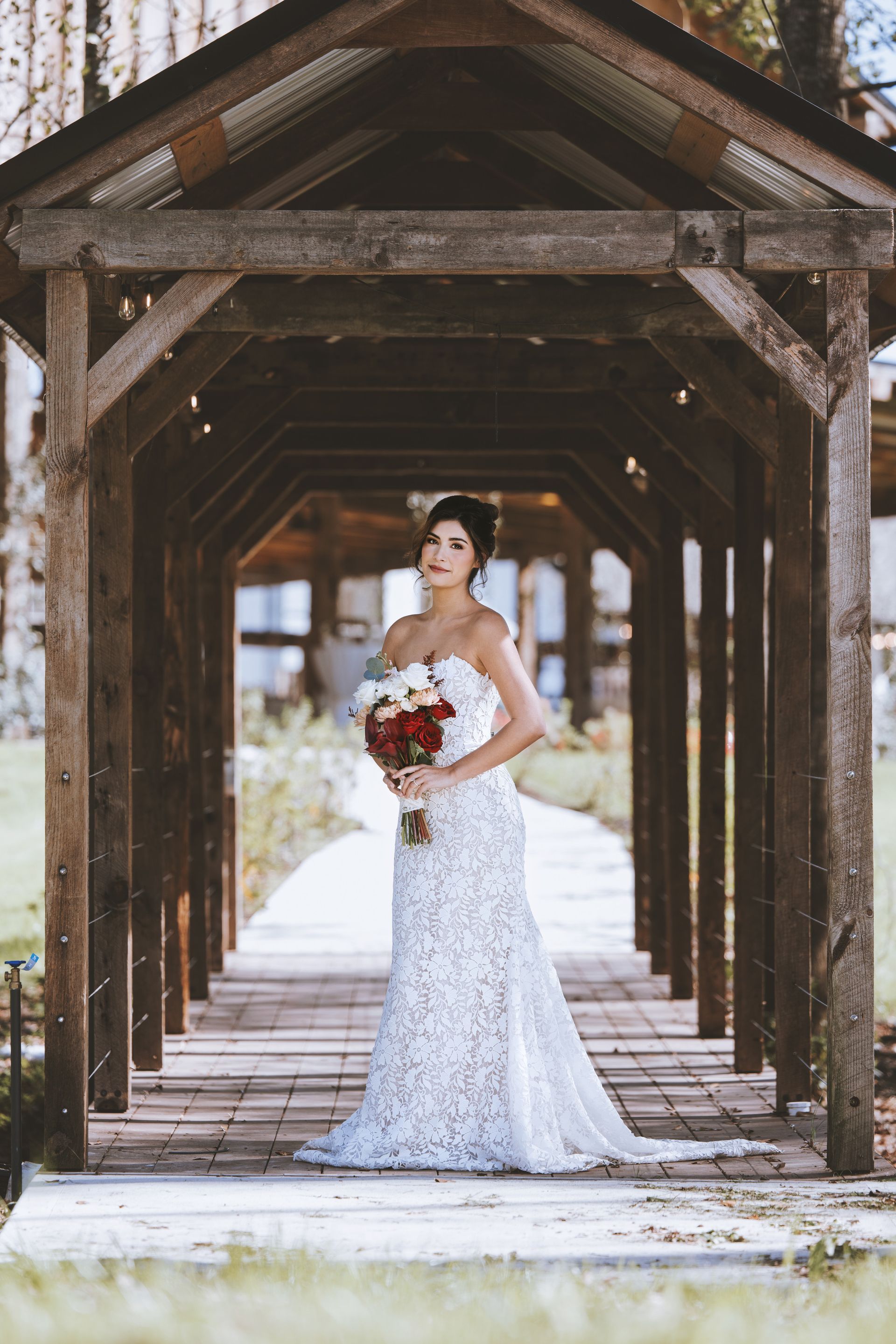Bride in a white lace gown holding a bouquet beneath a rustic wooden walkway outdoors
