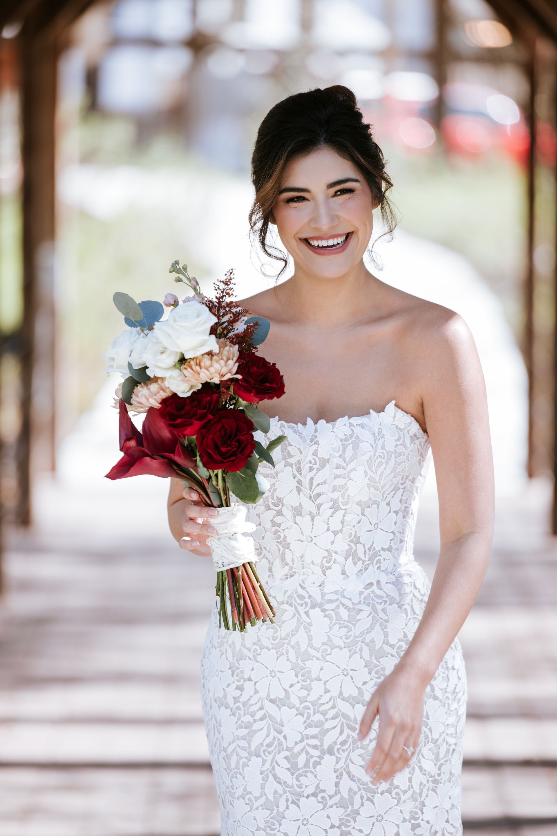 Bride in a strapless white gown holding a red-and-white bouquet, smiling on a sunlit walkway.
