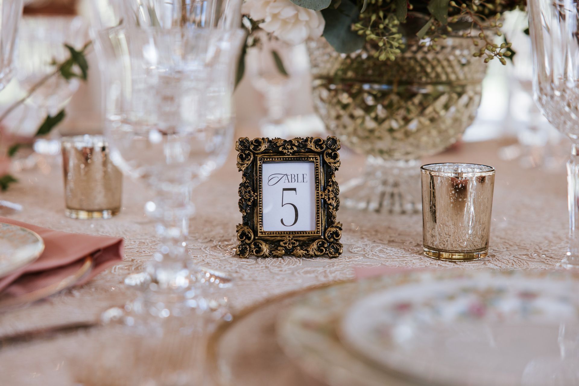 Table setting with a gold-framed table number 5 centerpiece, glassware, and soft floral decor