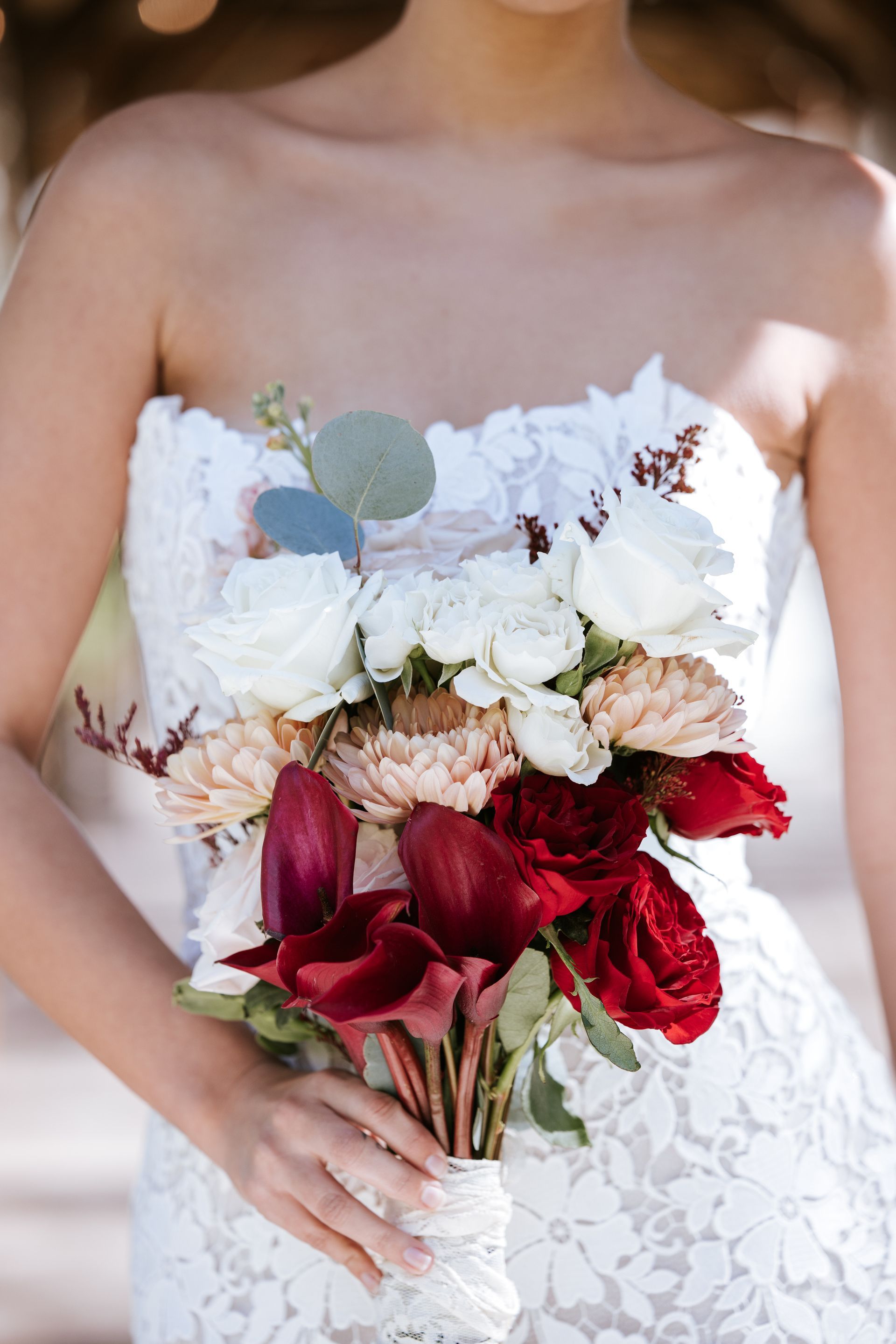 Bride in strapless white lace gown holding a bouquet of white, blush, and red flowers.