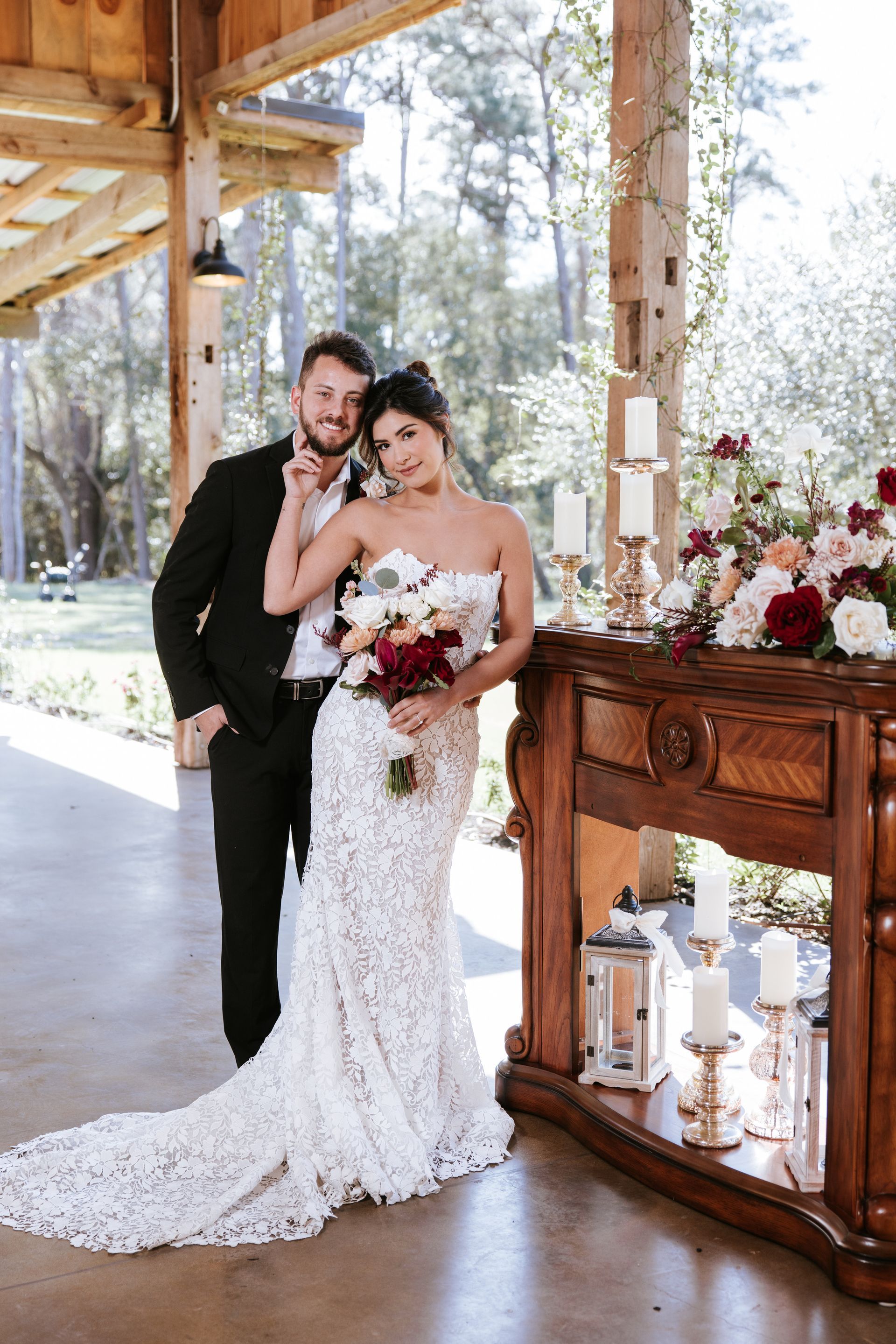 Bride and groom pose beside a rustic wooden table with candles and flowers under a covered outdoor pavilion.
