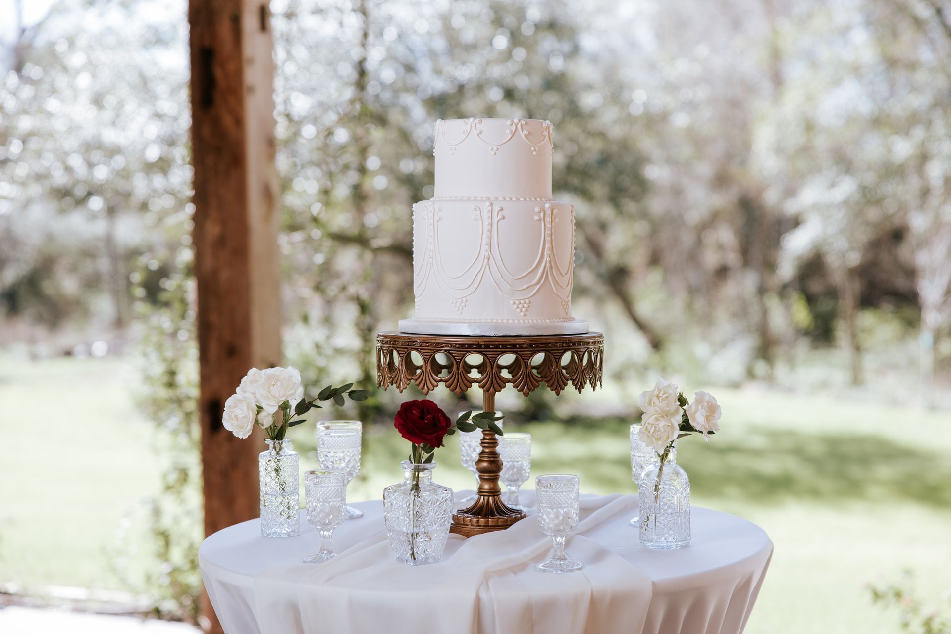 Tiered white wedding cake on a gold stand with flowers on a round table outdoors