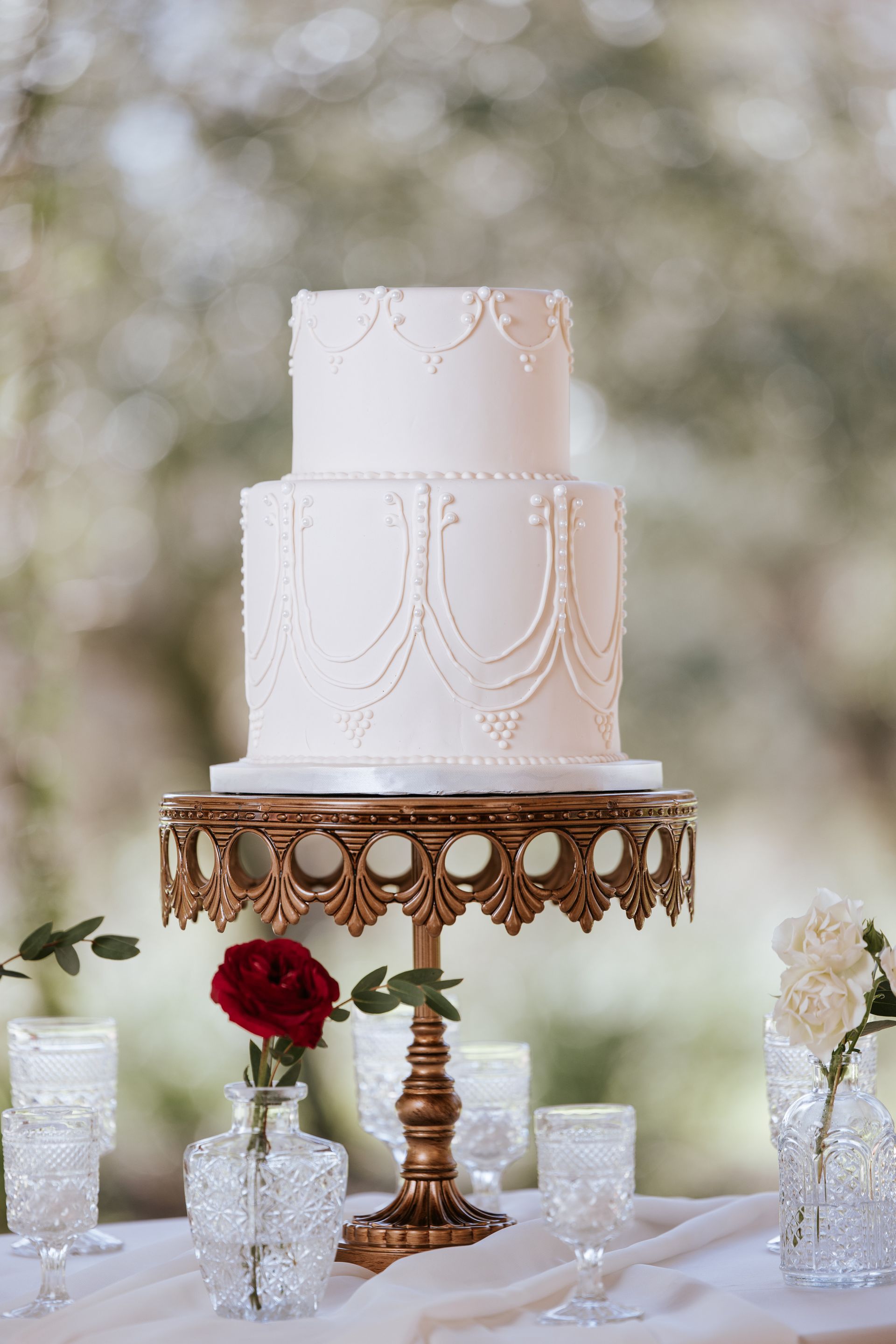 White tiered wedding cake on ornate gold stand with red rose and glass candle holders on a decorated table