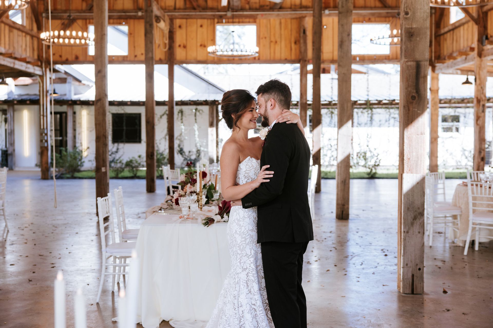 Bride and groom embracing in a rustic barn wedding venue, with a decorated table nearby.