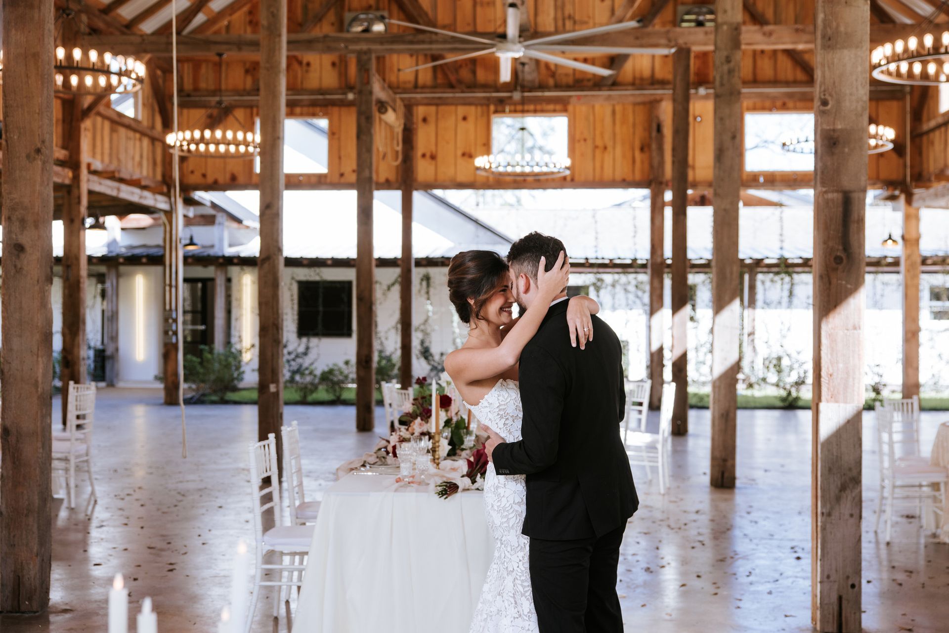 A bride and groom kiss in a rustic barn wedding venue with wooden beams and a decorated table nearby.