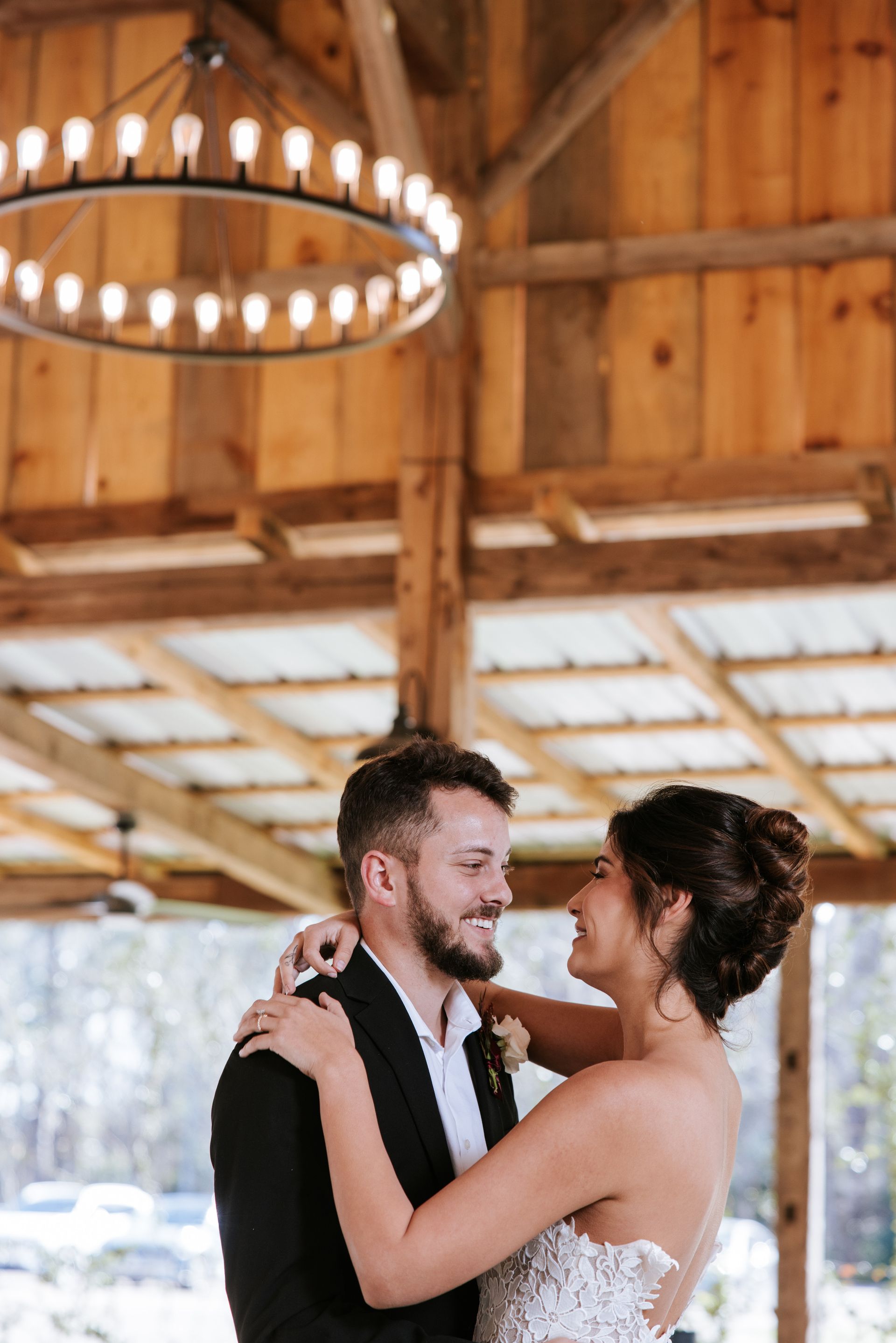 Couple embracing and smiling during a wedding dance under wooden rafters and a round chandelier