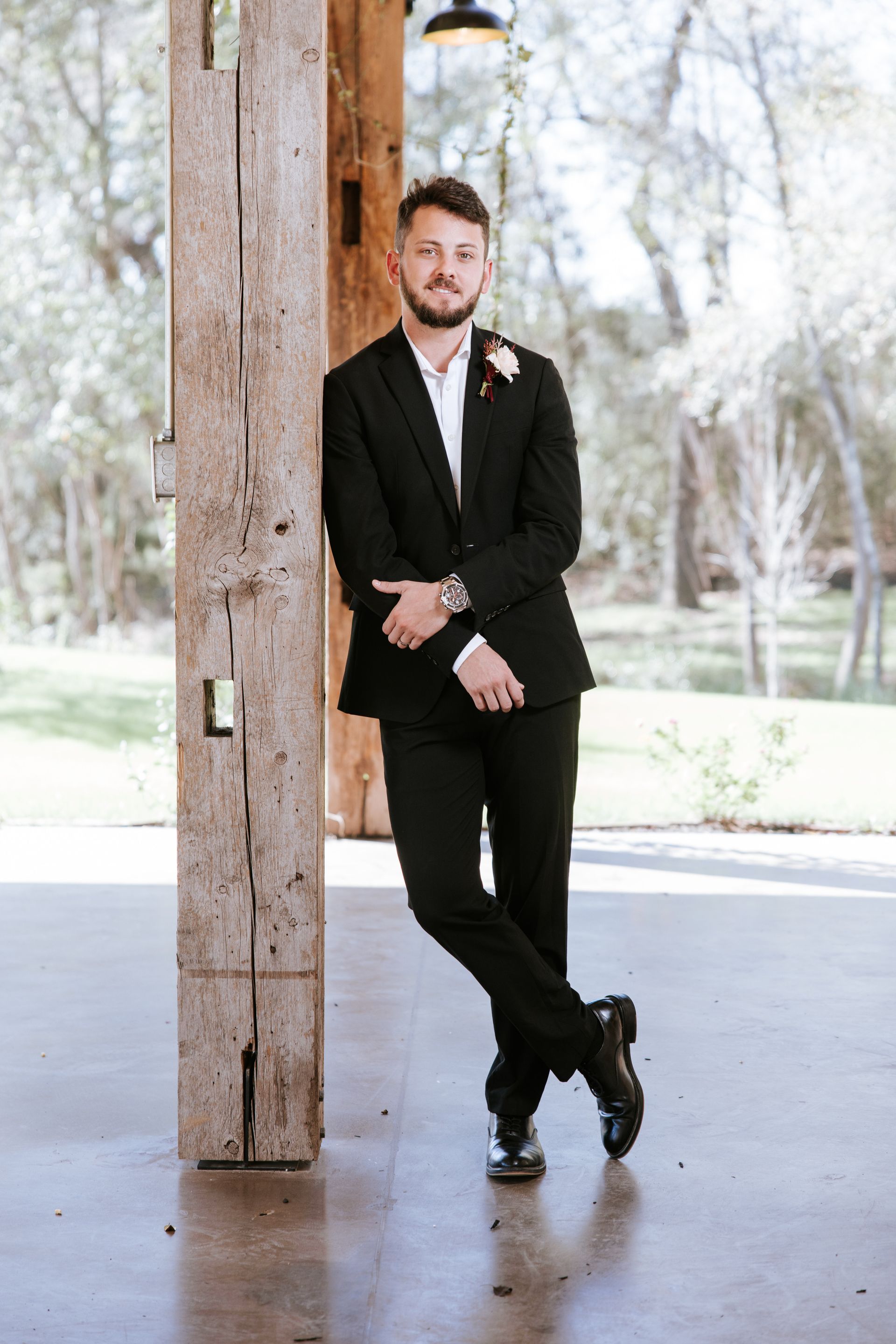 Man in a black suit leaning against a wooden post outdoors in a bright park setting