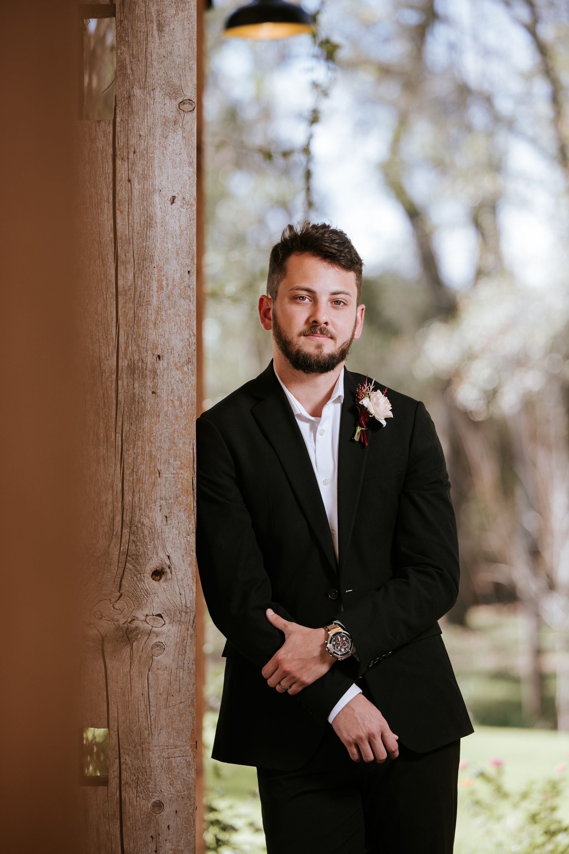 Man in black suit with boutonniere leaning against a wooden post outdoors