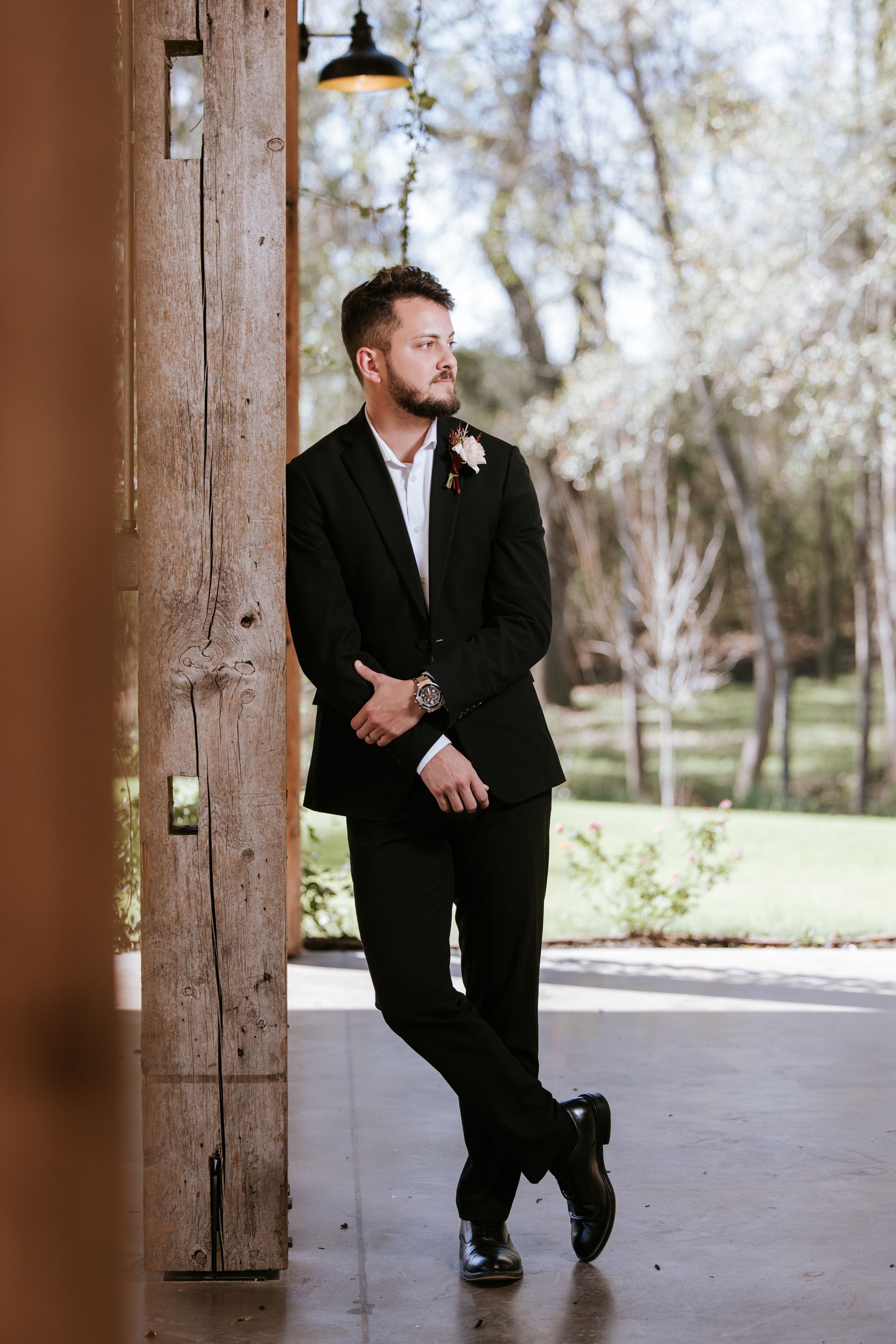 Man in a black suit leaning against a pillar outdoors on a sunny walkway