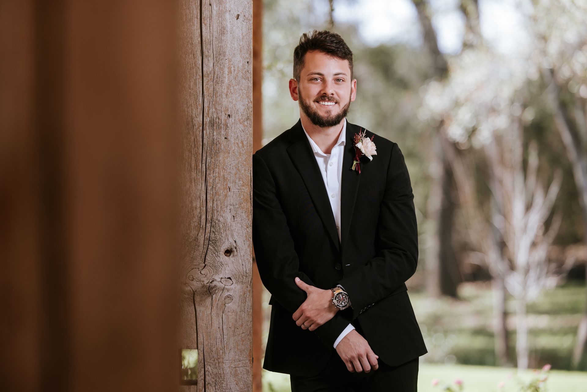 Man in a black suit standing by a wooden post outdoors, hands clasped, with a soft blurred background