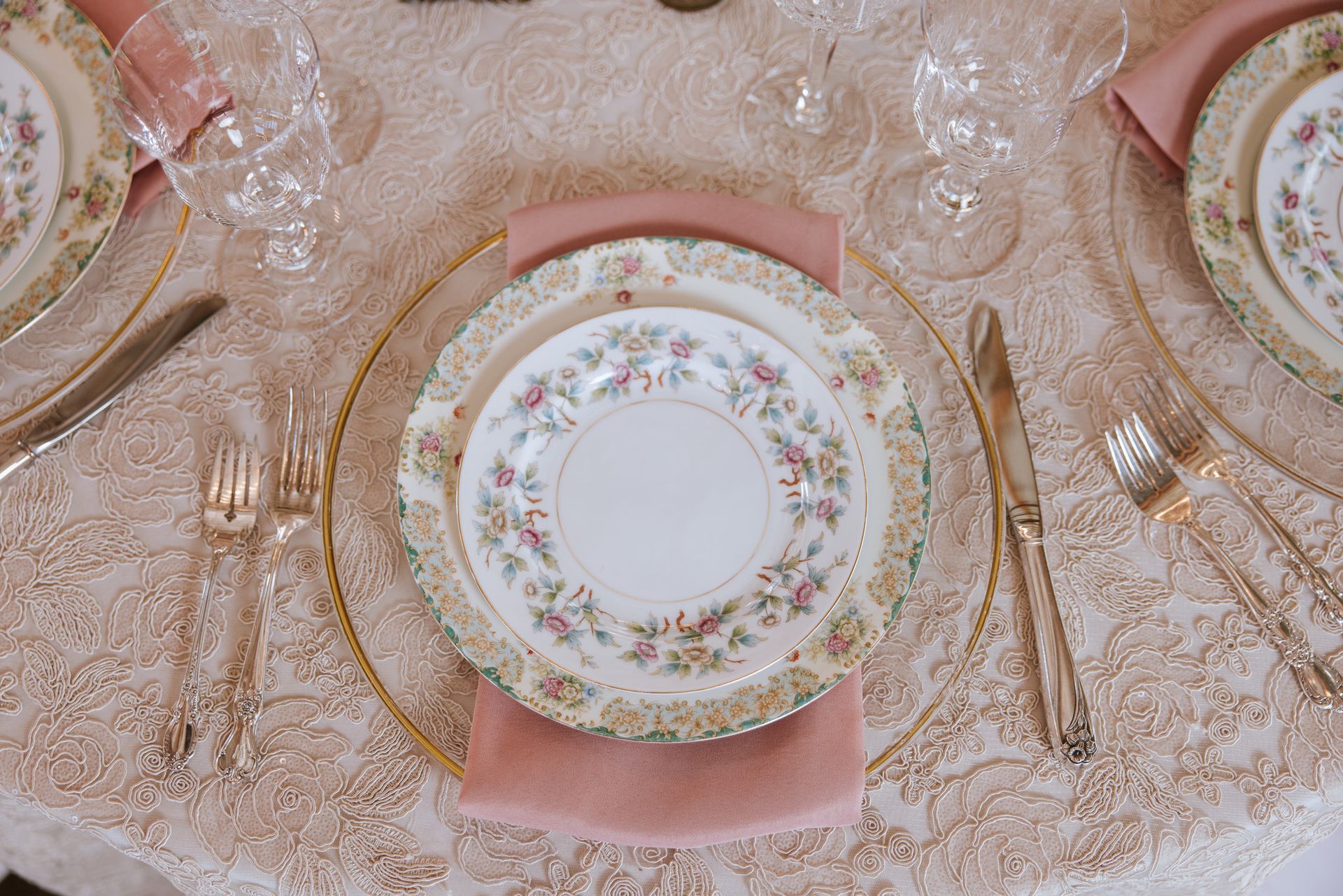 Elegant place setting with floral china, gold flatware, and pink napkins on a lace tablecloth