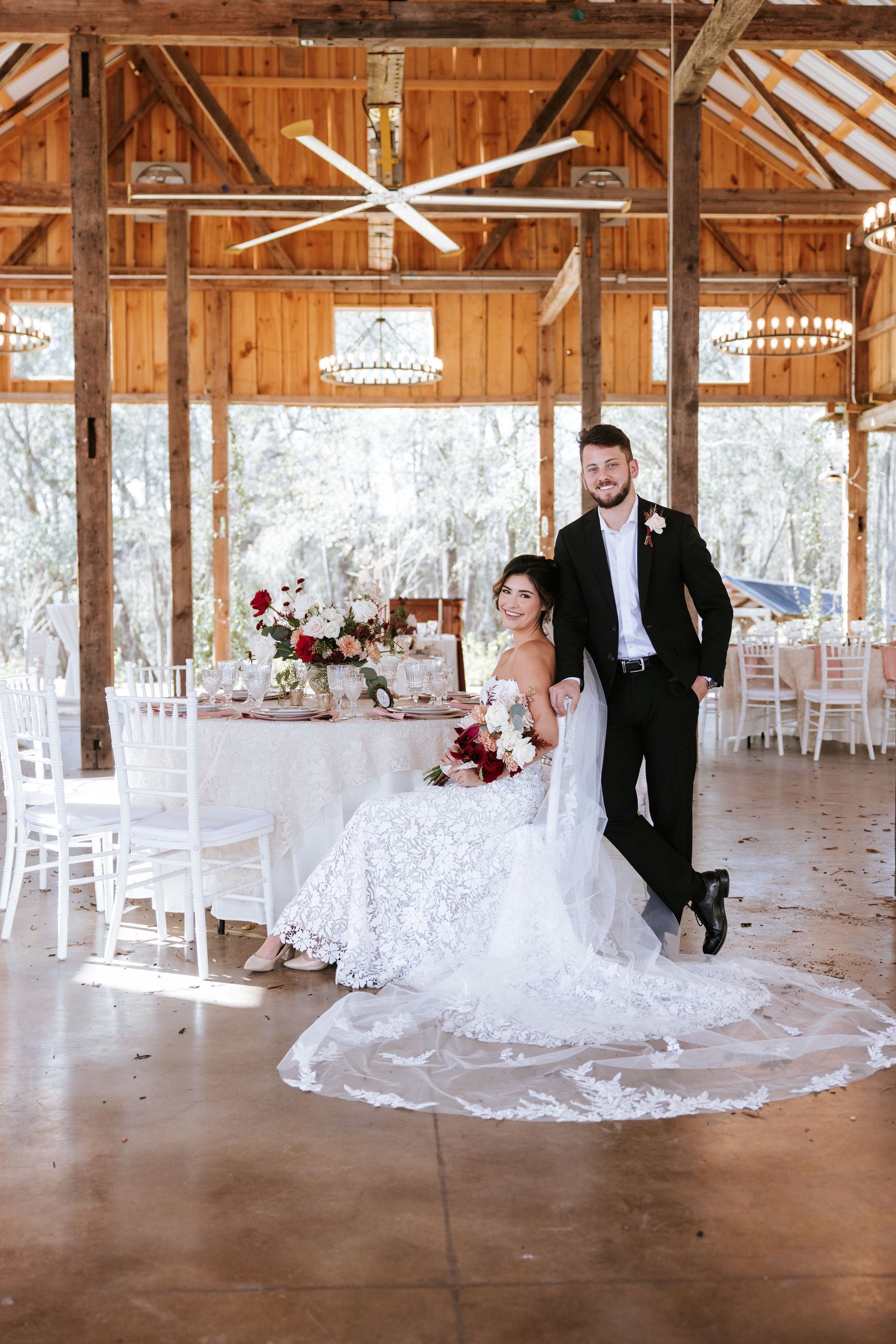 Bride and groom posing in a rustic wedding barn with white décor and a long table.
