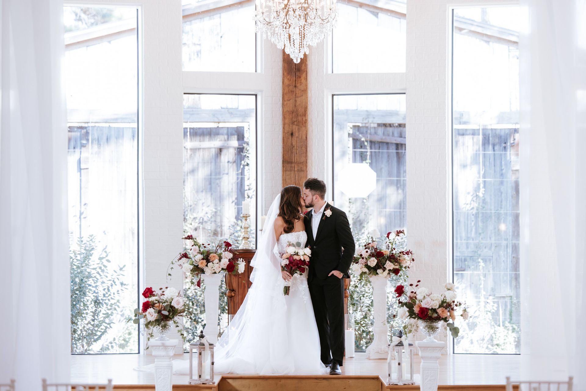 Bride and groom on a bright wedding stage with white floral decor and tall windows