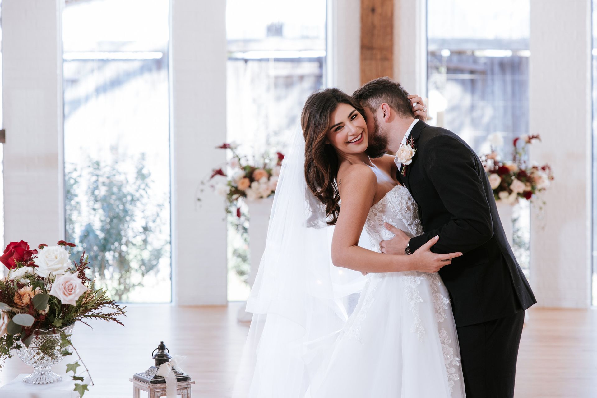 Bride and groom embrace and smile in a bright wedding venue with floral arrangements.
