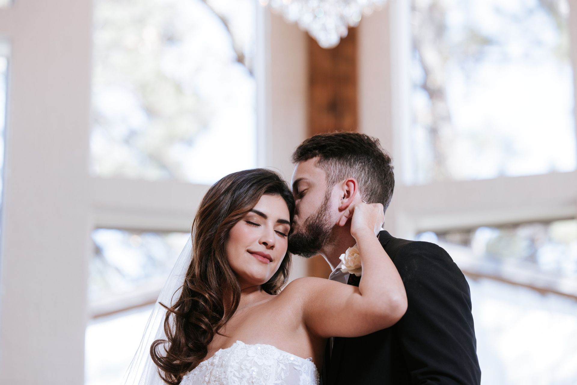 Bride and groom embrace in a bright church, with the groom kissing her cheek.