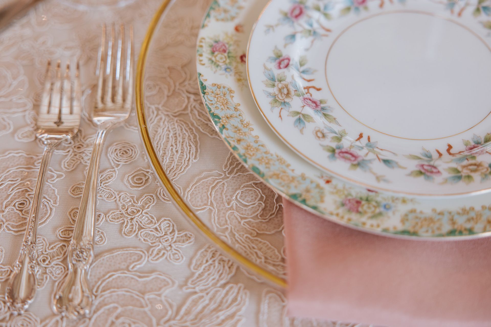 Elegant table setting with floral china, gold-rimmed plates, crystal glasses, and a pink napkin
