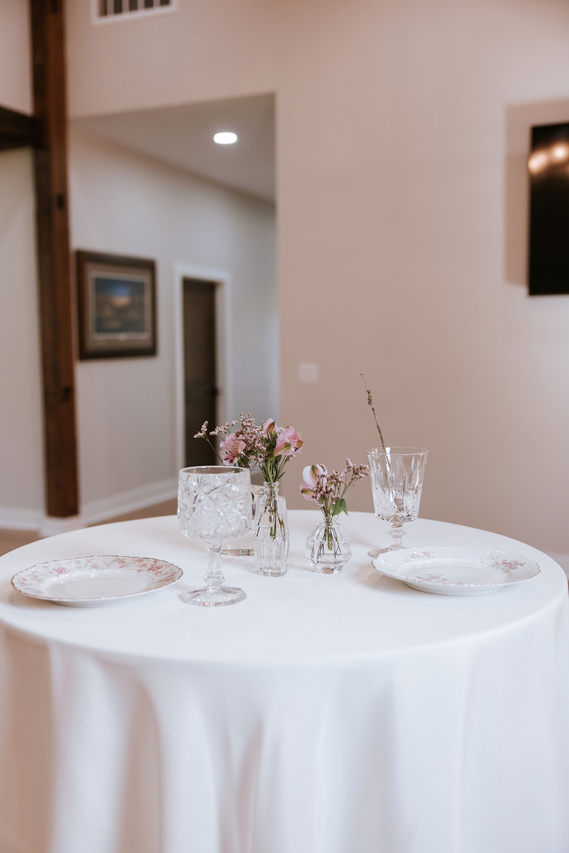 Round white-draped table set with plates, glasses, and floral centerpieces in a bright room
