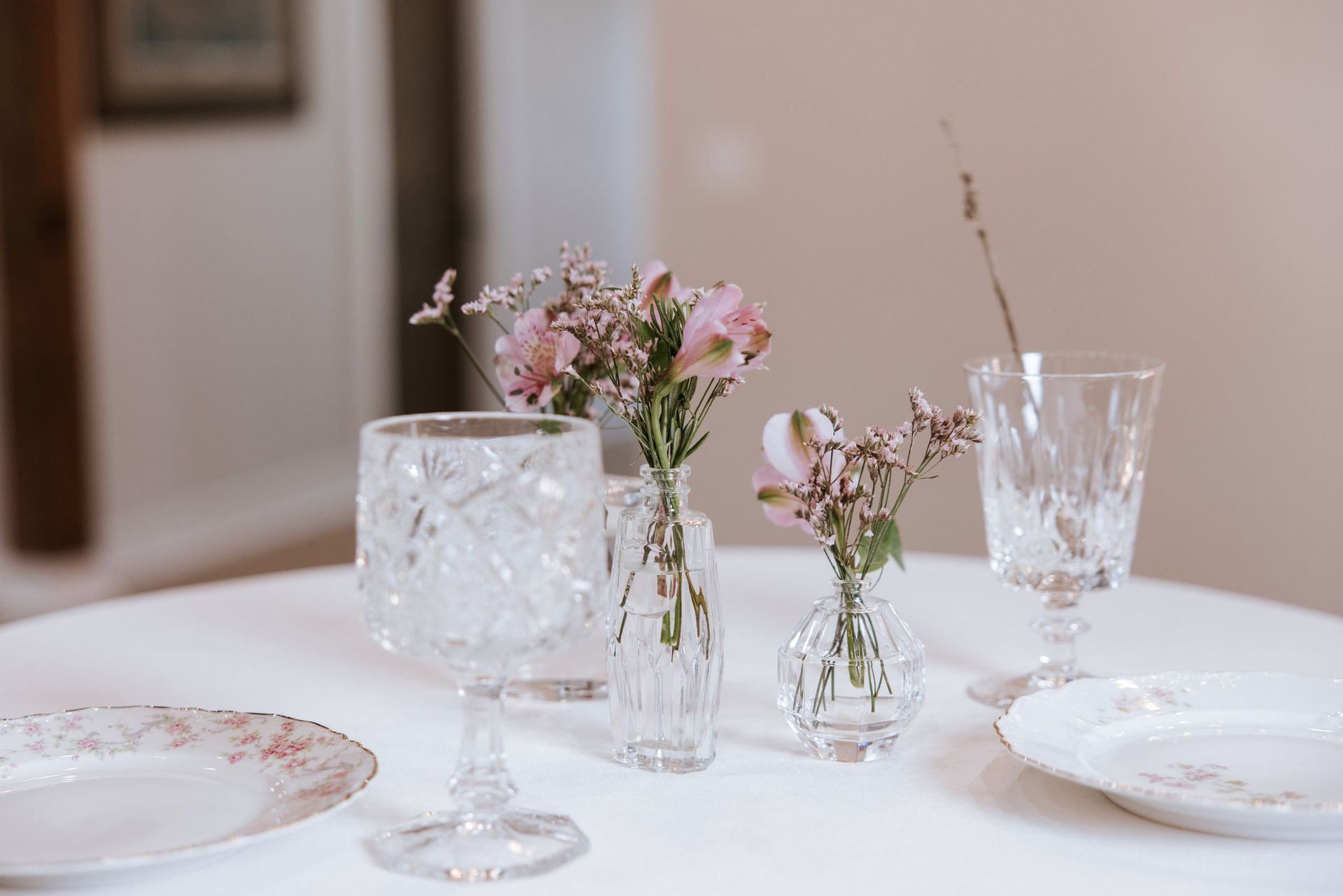 Elegant table setting with crystal glassware and small pink floral arrangements on a white tablecloth